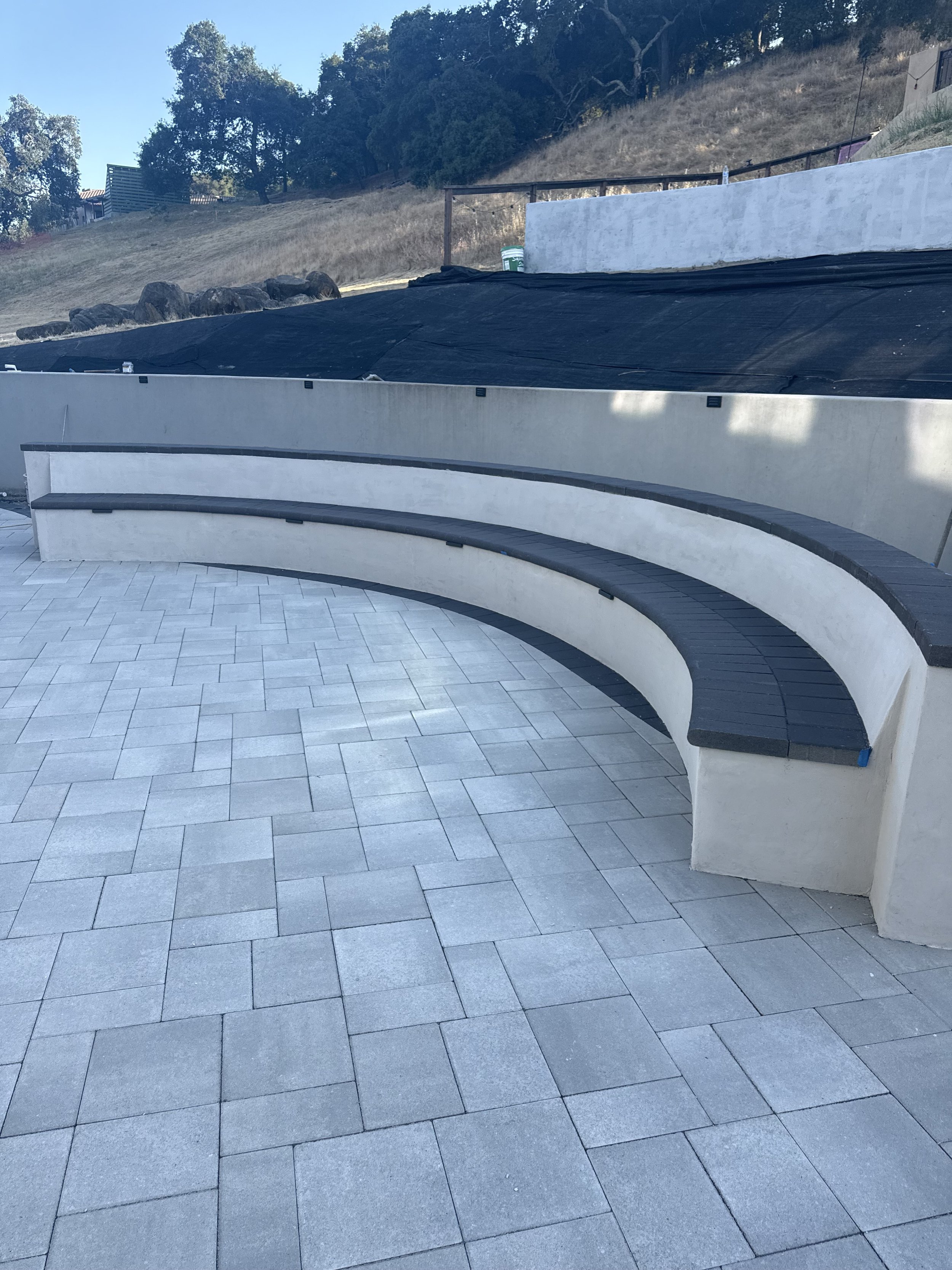 Curved outdoor seating area with white concrete benches and black tiled surface, overlooking a hill with dry grass and trees, under a clear blue sky.