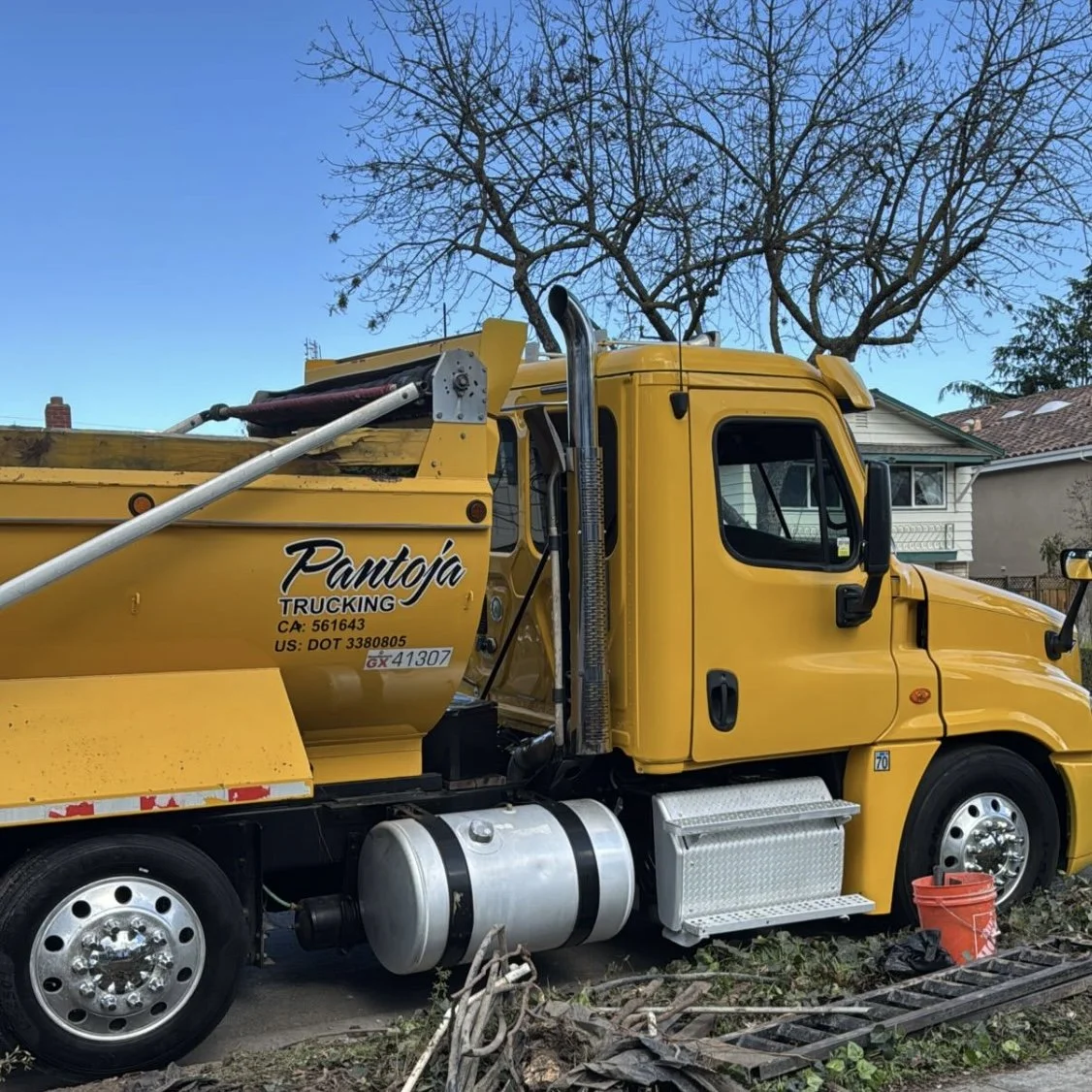 Yellow utility truck parked in front of a house with a leafless tree in the background. The truck has the words 'Pantoja Trucking' on the side and various equipment attached.