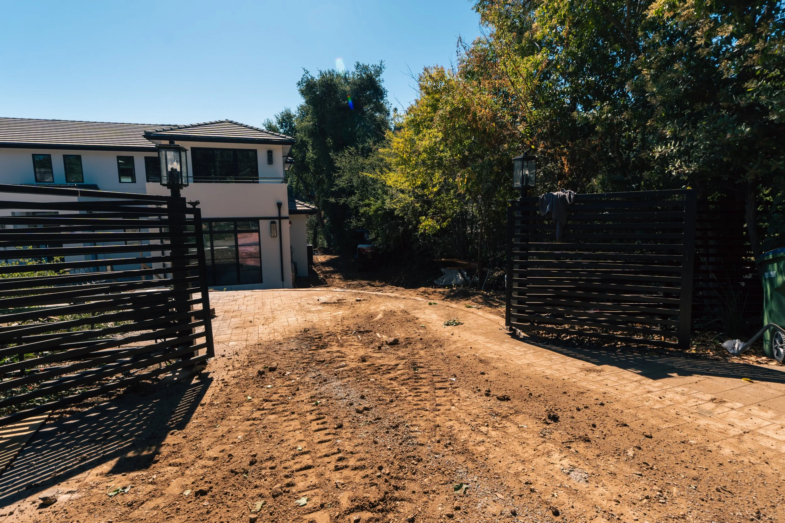 A modern white house with a tiled roof and black window frames, located behind a brown dirt driveway with tire tracks, flanked by black metal gates, and surrounded by green trees under a clear blue sky.