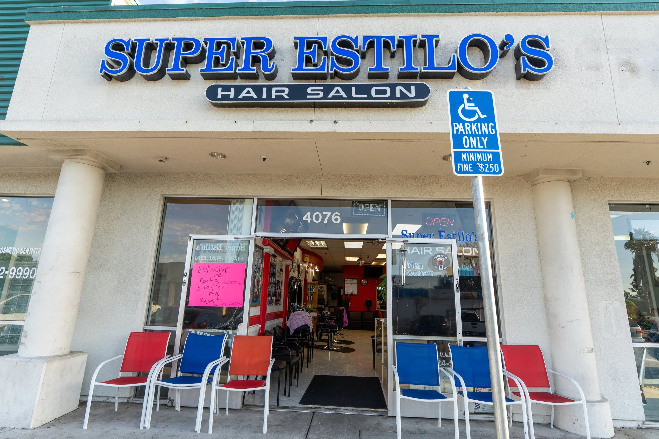 Exterior view of Super Estilo's Hair Salon with a blue and white sign, a handicapped parking sign, red and blue chairs outside, and the salon entrance with open doors showing hairdressing chairs inside.