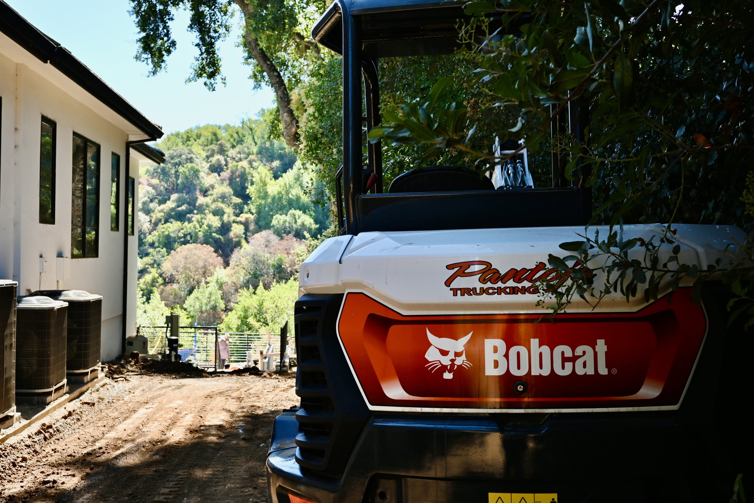 A Bobcat brand construction or landscaping vehicle parked on a dirt path next to a white building, with a background of green trees and hills under a sunny sky.