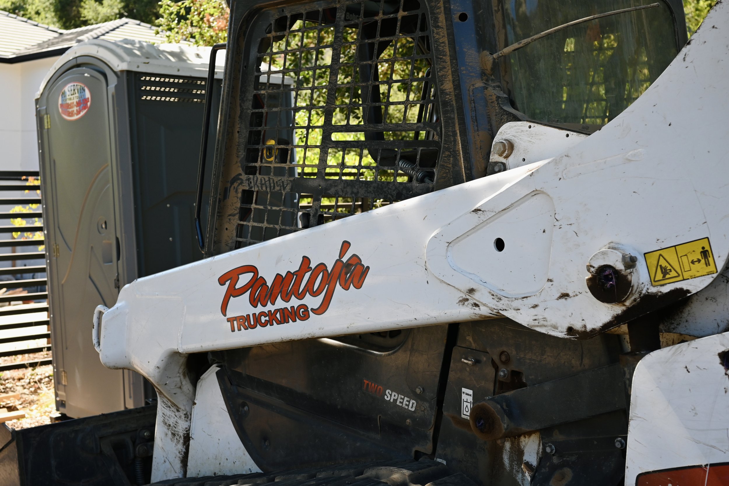Close-up of a white and black skid steer loader with a yellow warning sticker, dirt, and the logo 'Pantoja Trucking' on the side, parked outdoors.