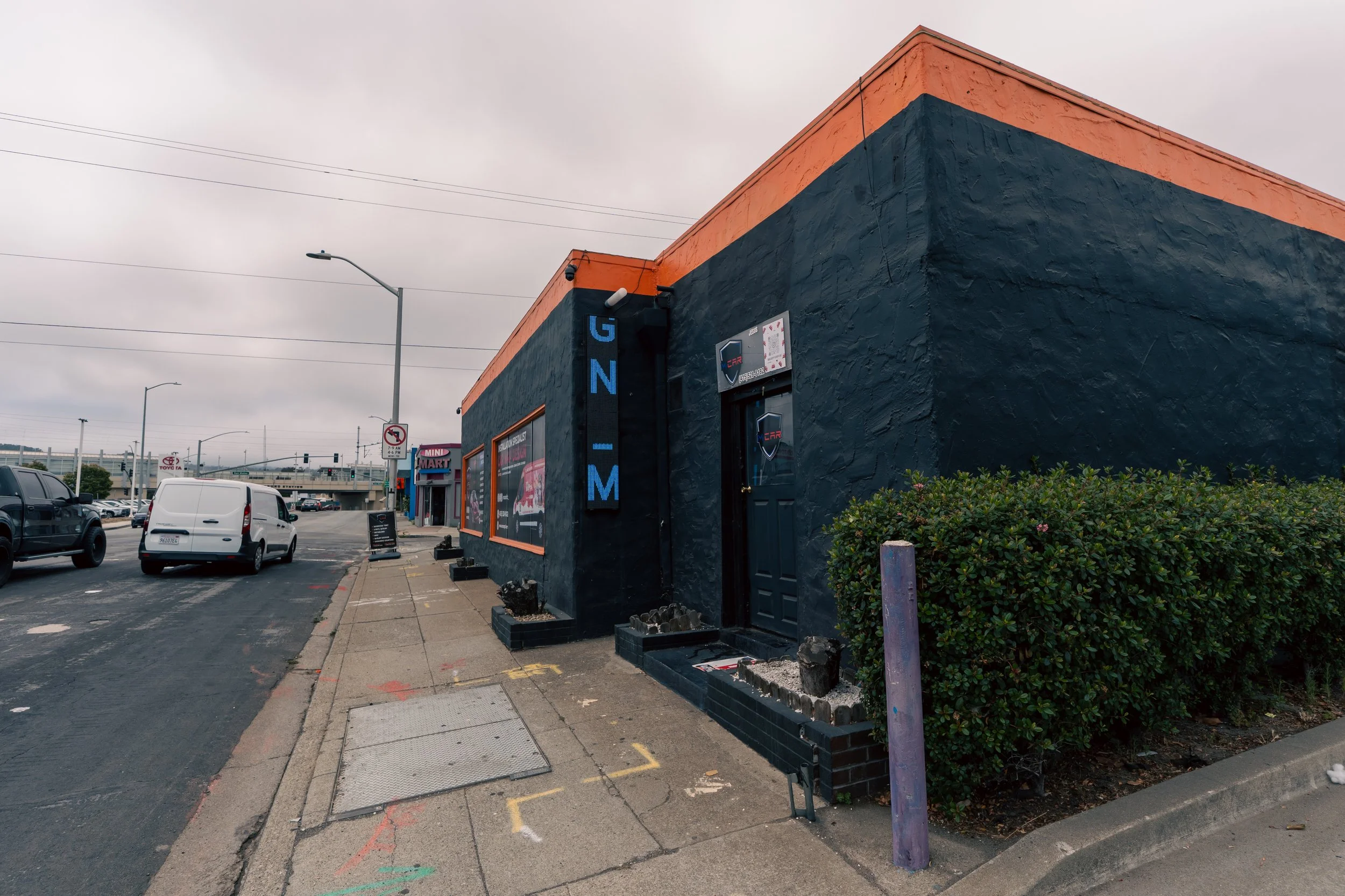 A street view with a black building featuring blue and orange accents, and a sign reading 'G N I M' on the corner. The sidewalk in front has small planters and debris, with cars parked along the street and an overpass in the background.