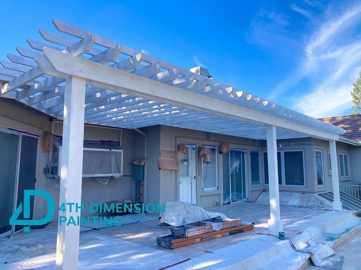Construction of a white pergola structure over a house patio with cloudy blue sky in the background.