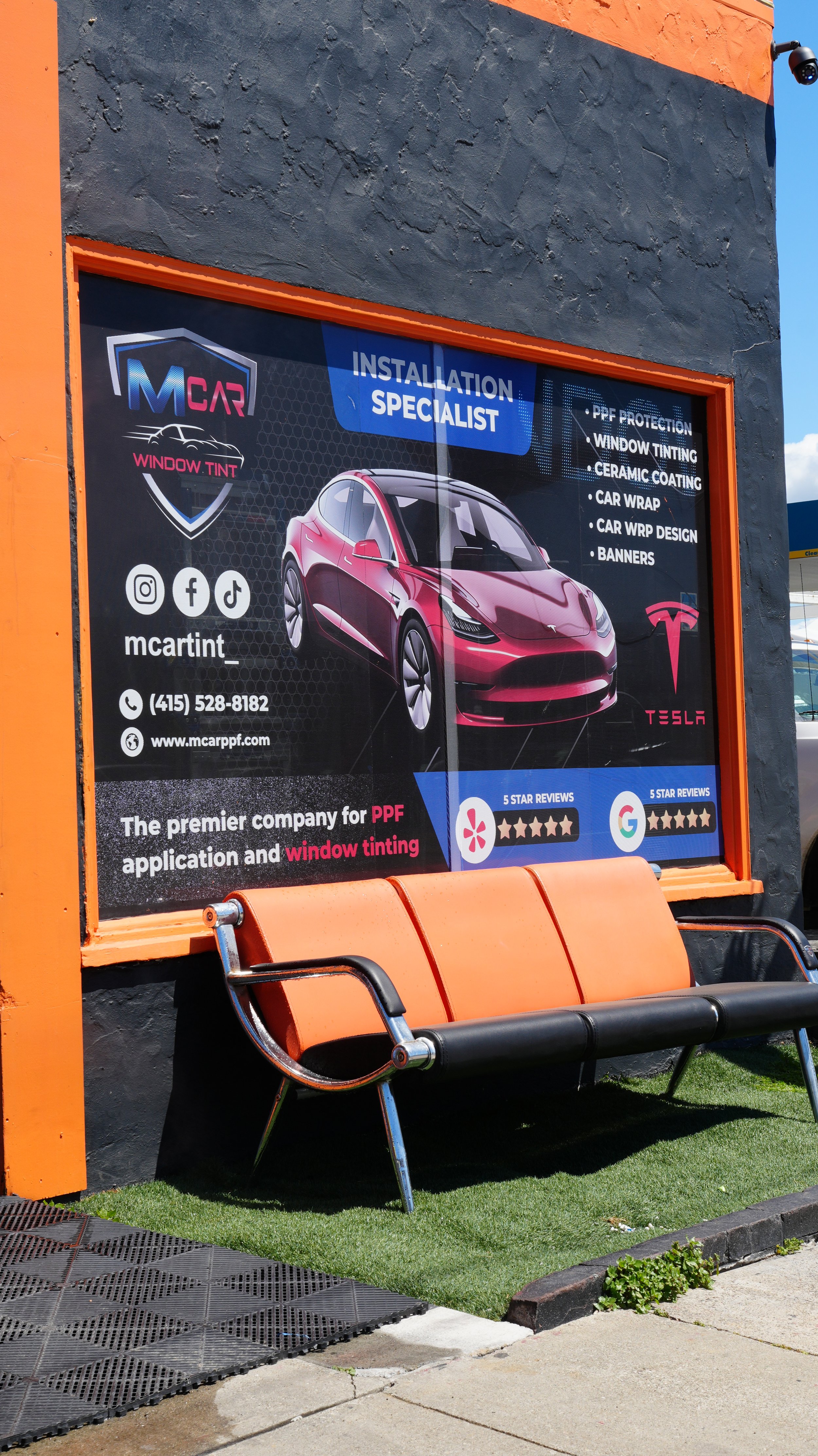 Sign in front of a building advertising window tinting services with a picture of a red Tesla car, social media icons, and contact information. A bench is placed below the sign.