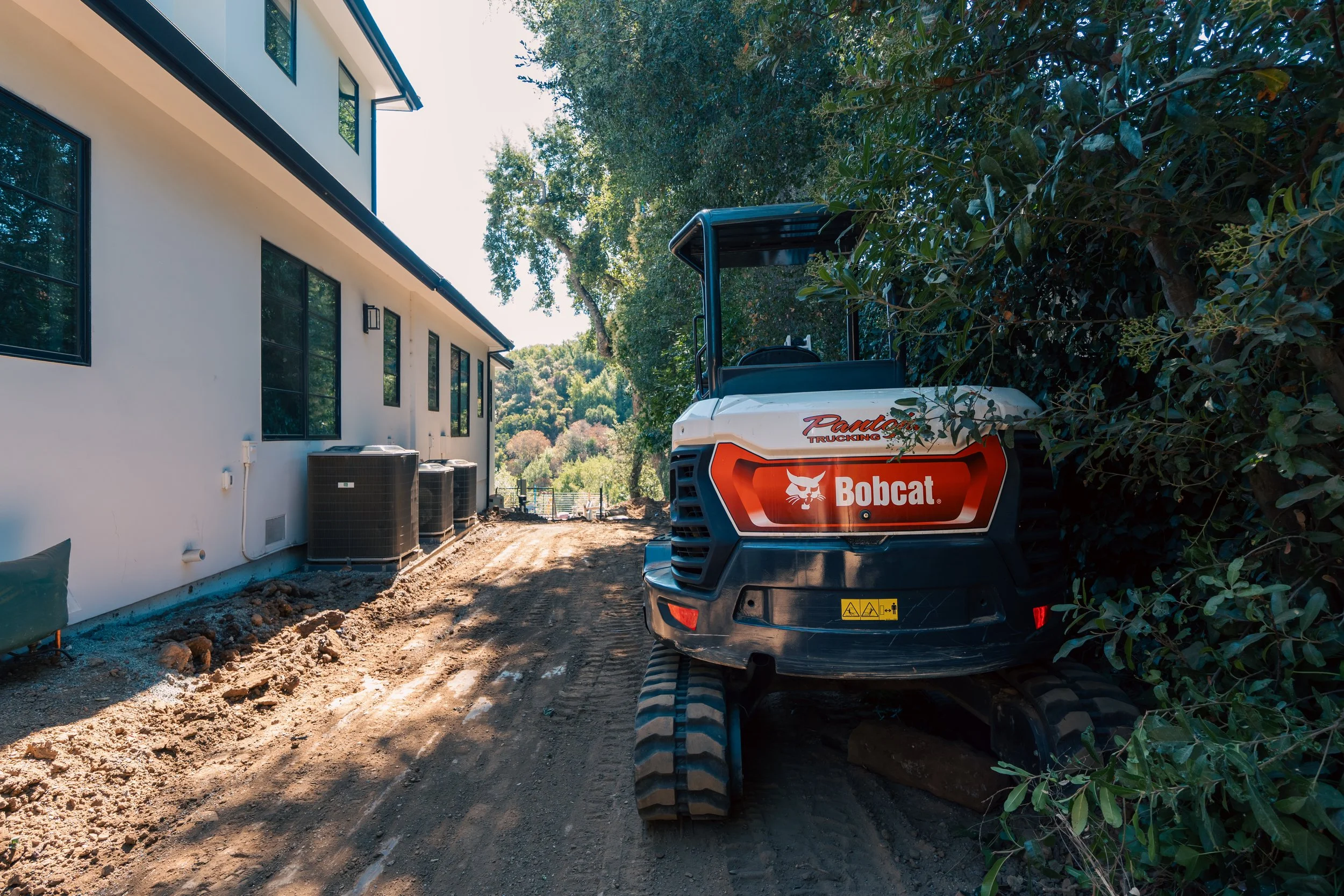 Construction site with a white and black Bobcat mini excavator next to a white house with black window frames, on a dirt pathway surrounded by trees and green foliage.