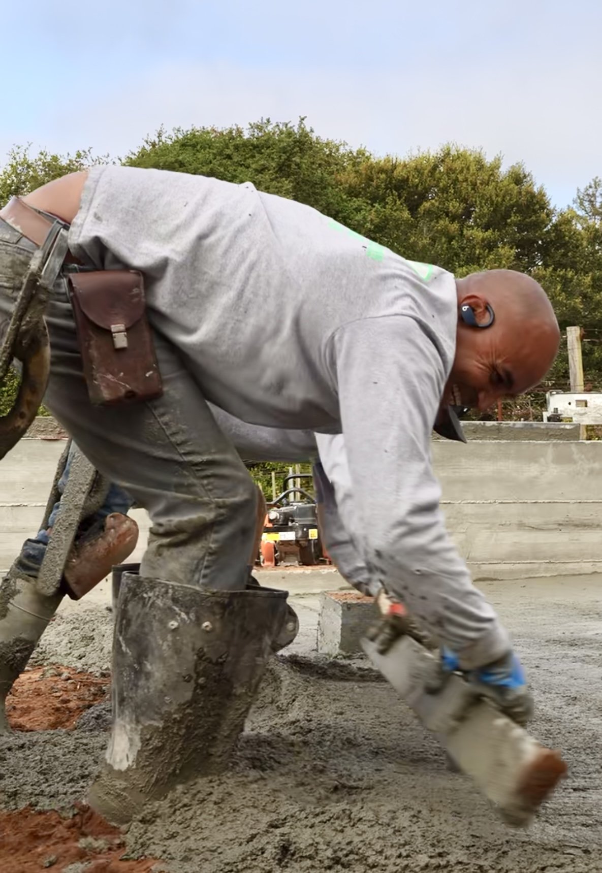 A construction worker in gray clothing and boots, wearing a headset, is spreading wet cement on a construction site.