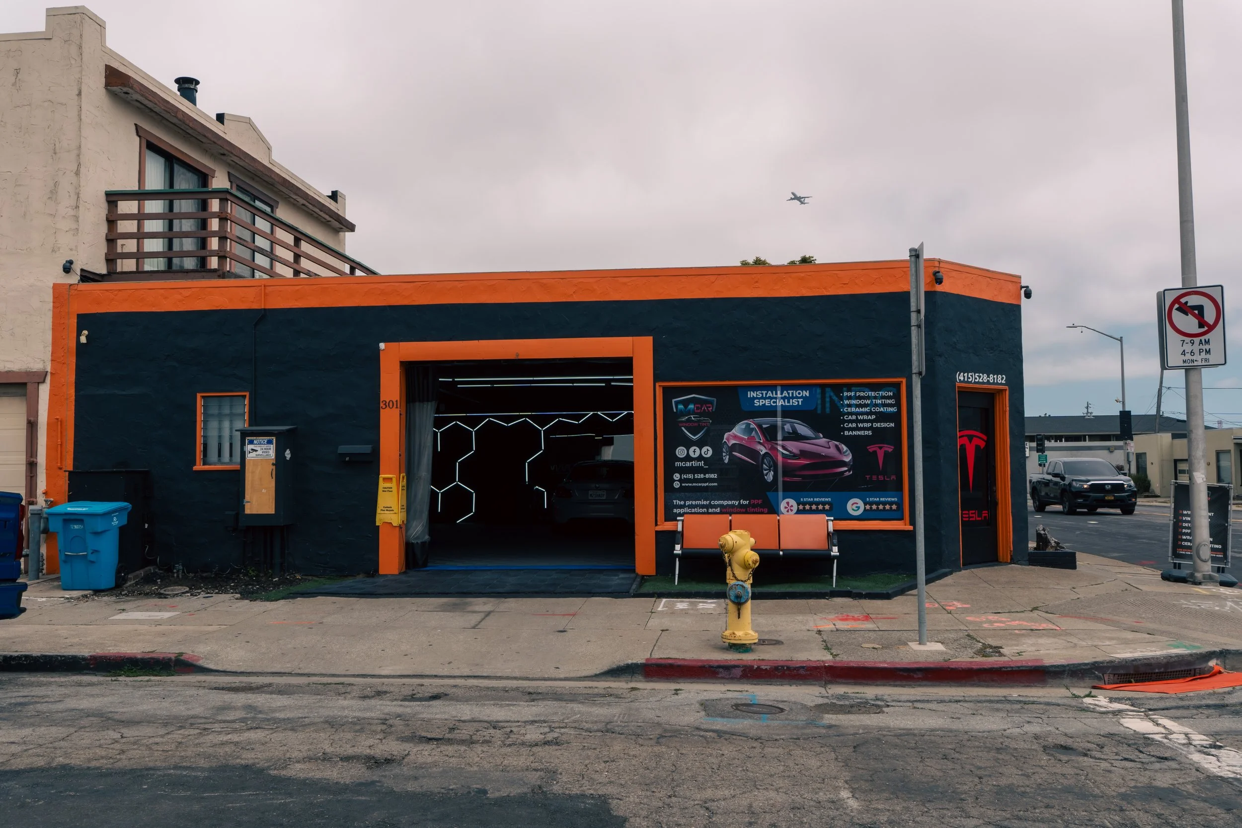 A black building with orange trim housing a Tesla window tinting and car wrapping shop. There is a large sign displaying Tesla vehicles and services on the front, with a Tesla logo on a door to the right. A parking garage with cars inside is visible 