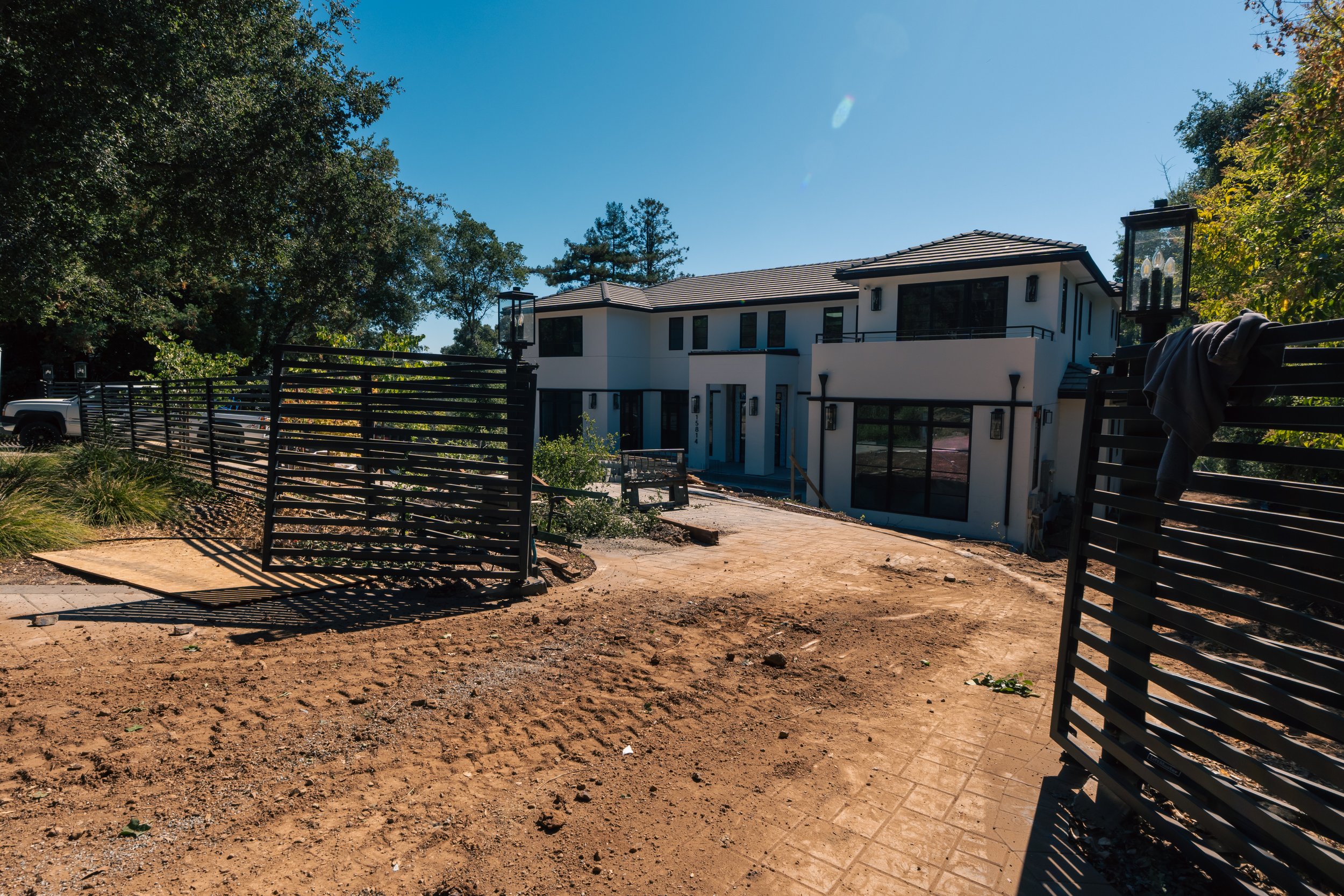 Construction site with new modern house, wooden pathways, and black fencing, surrounded by trees under a clear blue sky.
