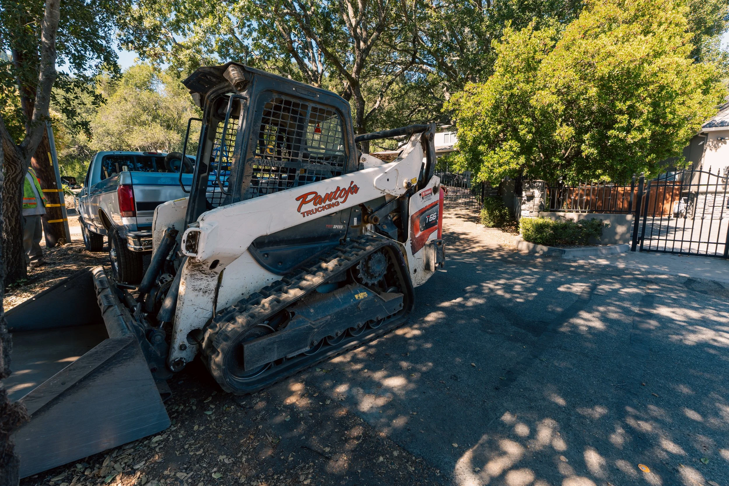 A small construction vehicle, likely a skid steer loader, parked on a street next to a curb with trees and a house in the background.