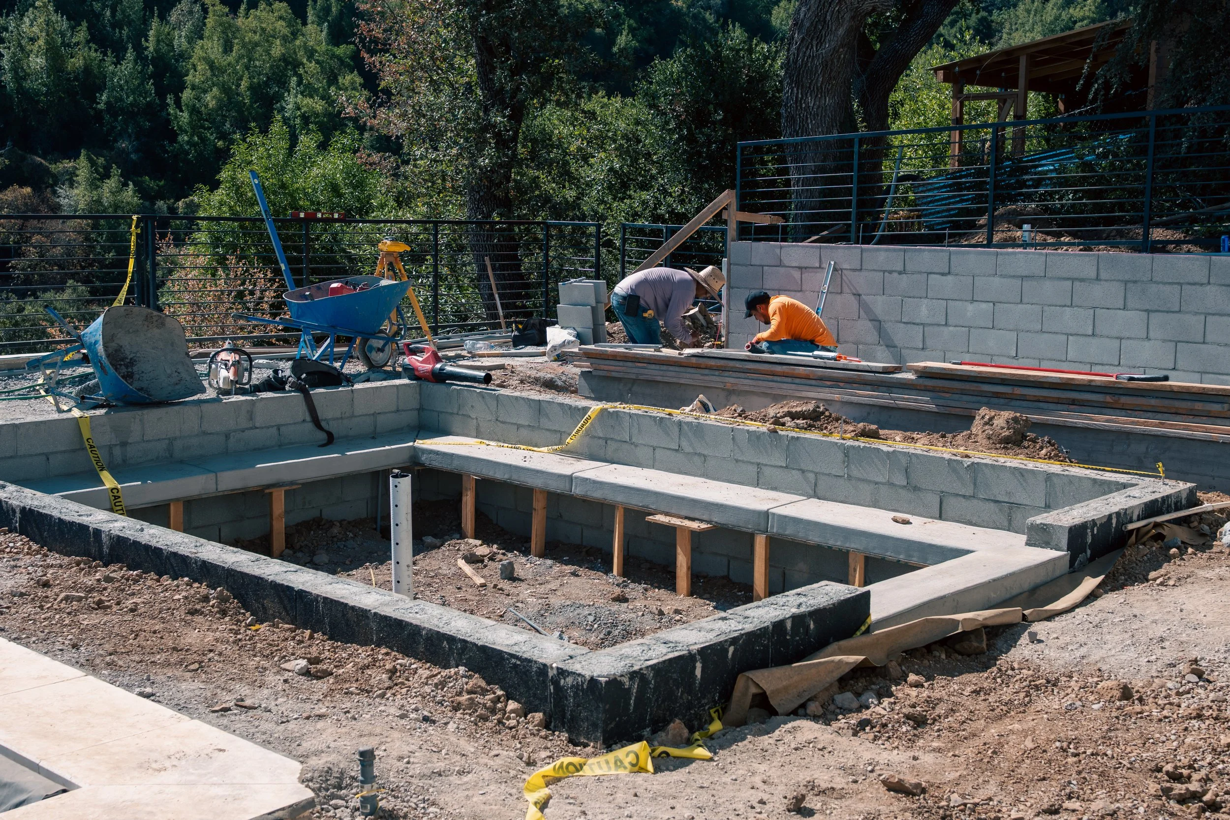 Construction workers building a swimming pool with cinder blocks and concrete, tools and construction materials scattered around, in a backyard with trees and a fence.