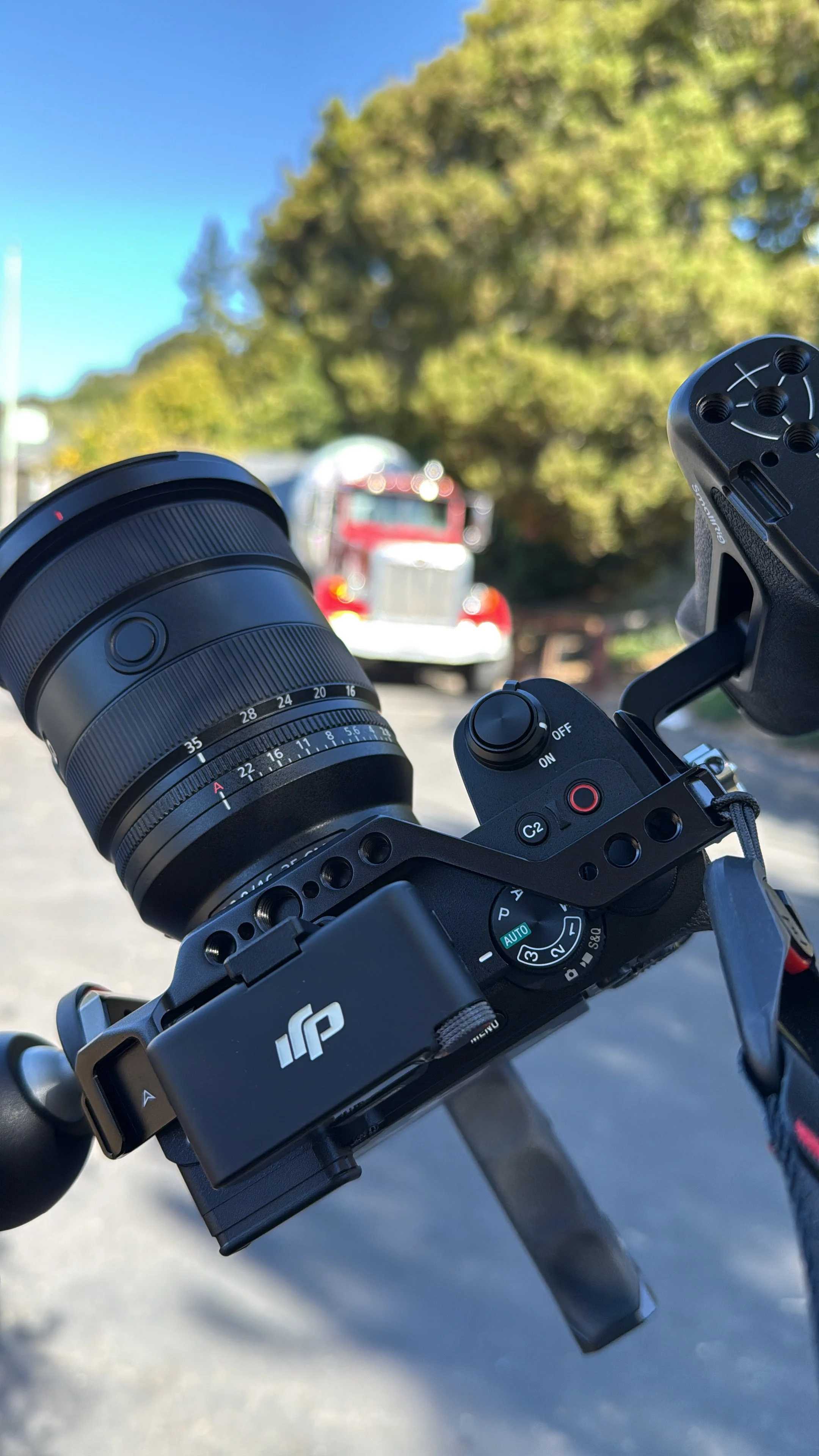 Camera mounted on a tripod, with a blurred red and white truck in background and trees behind.