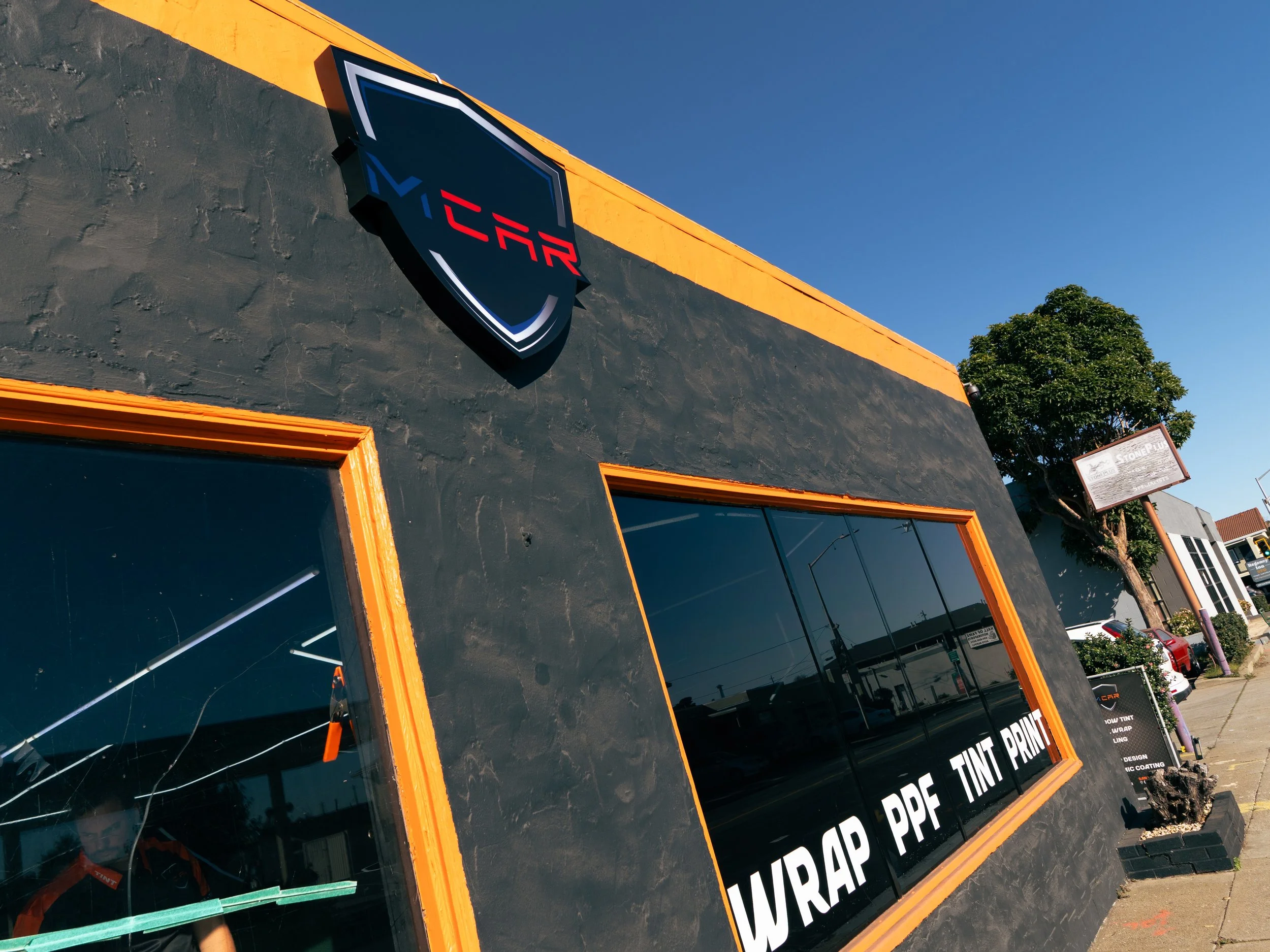 Black building with orange trim and a logo of a shield with the letters 'ICAR' on it, and a window displaying many vinyl wrap and printing services listed.
