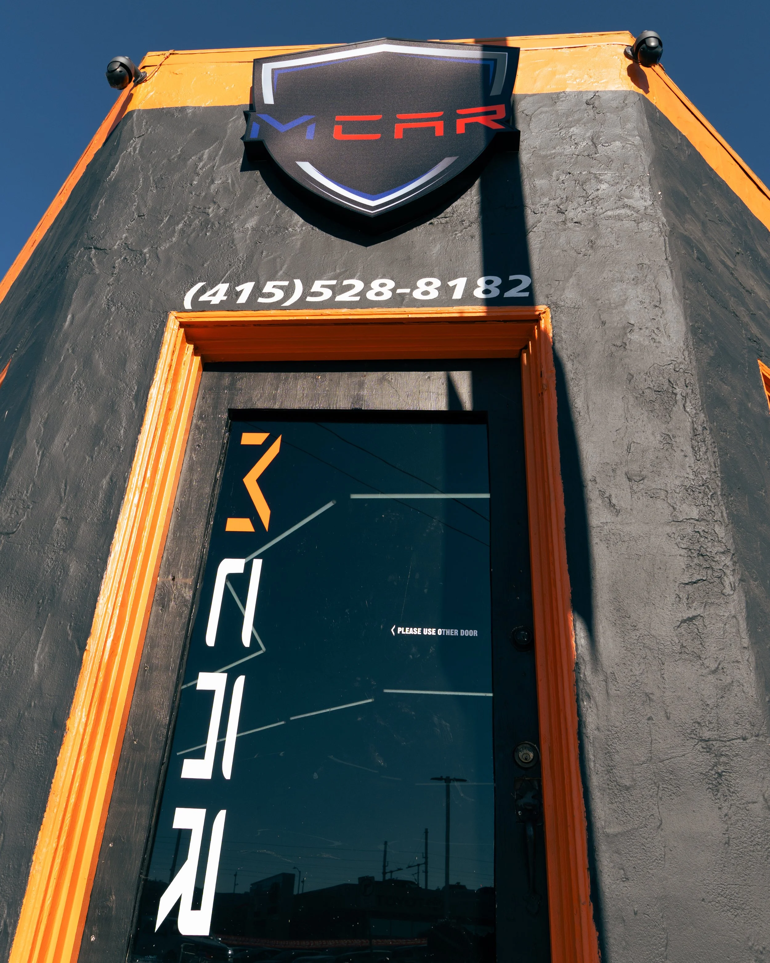 Exterior view of a building with black textured walls and orange trim, featuring a black door with white and orange graphics, a sign for MYCAR with a shield-shaped logo, and a phone number (415) 528-8182.