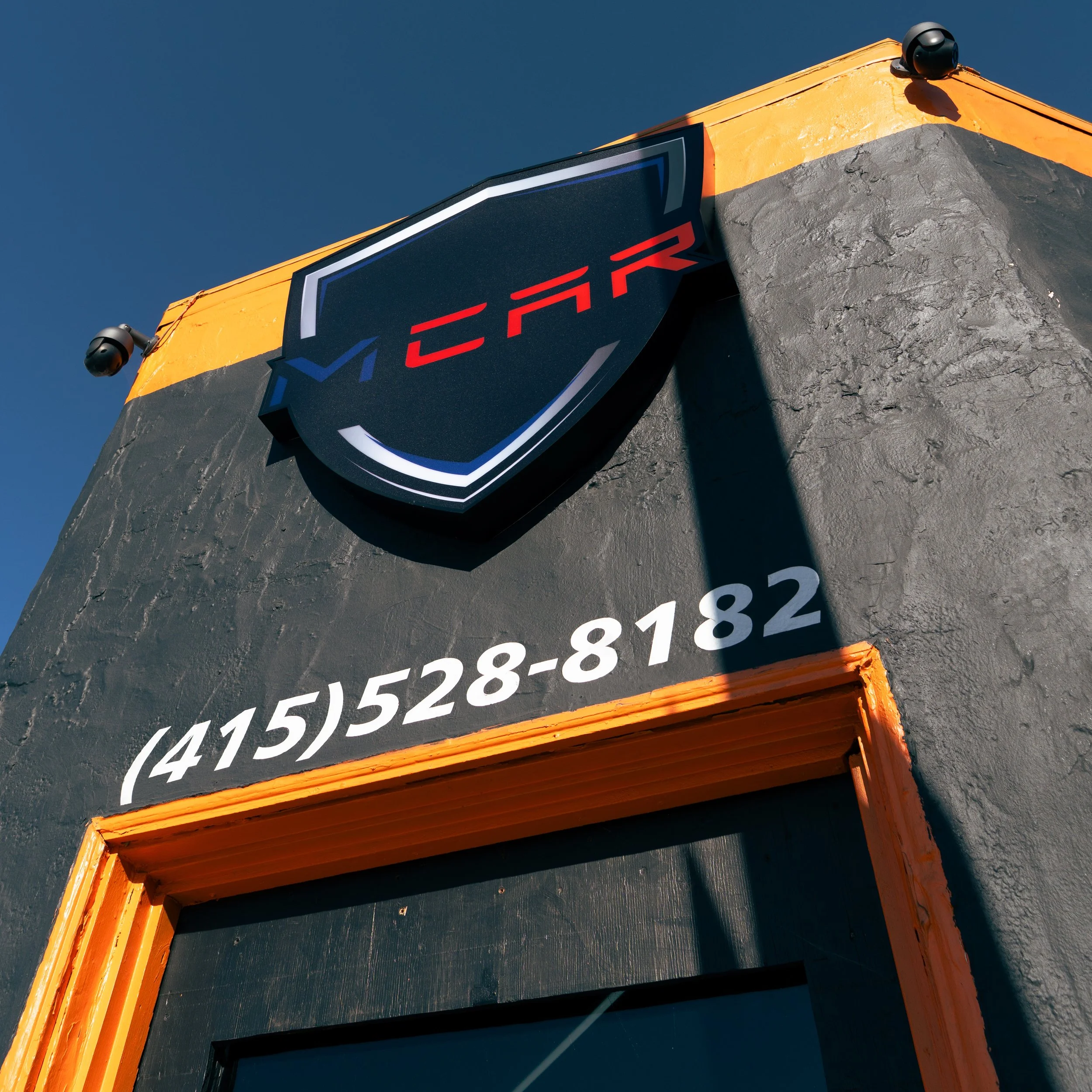 Building exterior with a black wall, orange trim, and a shield-shaped sign that has a red and blue logo, as well as a phone number in white text.