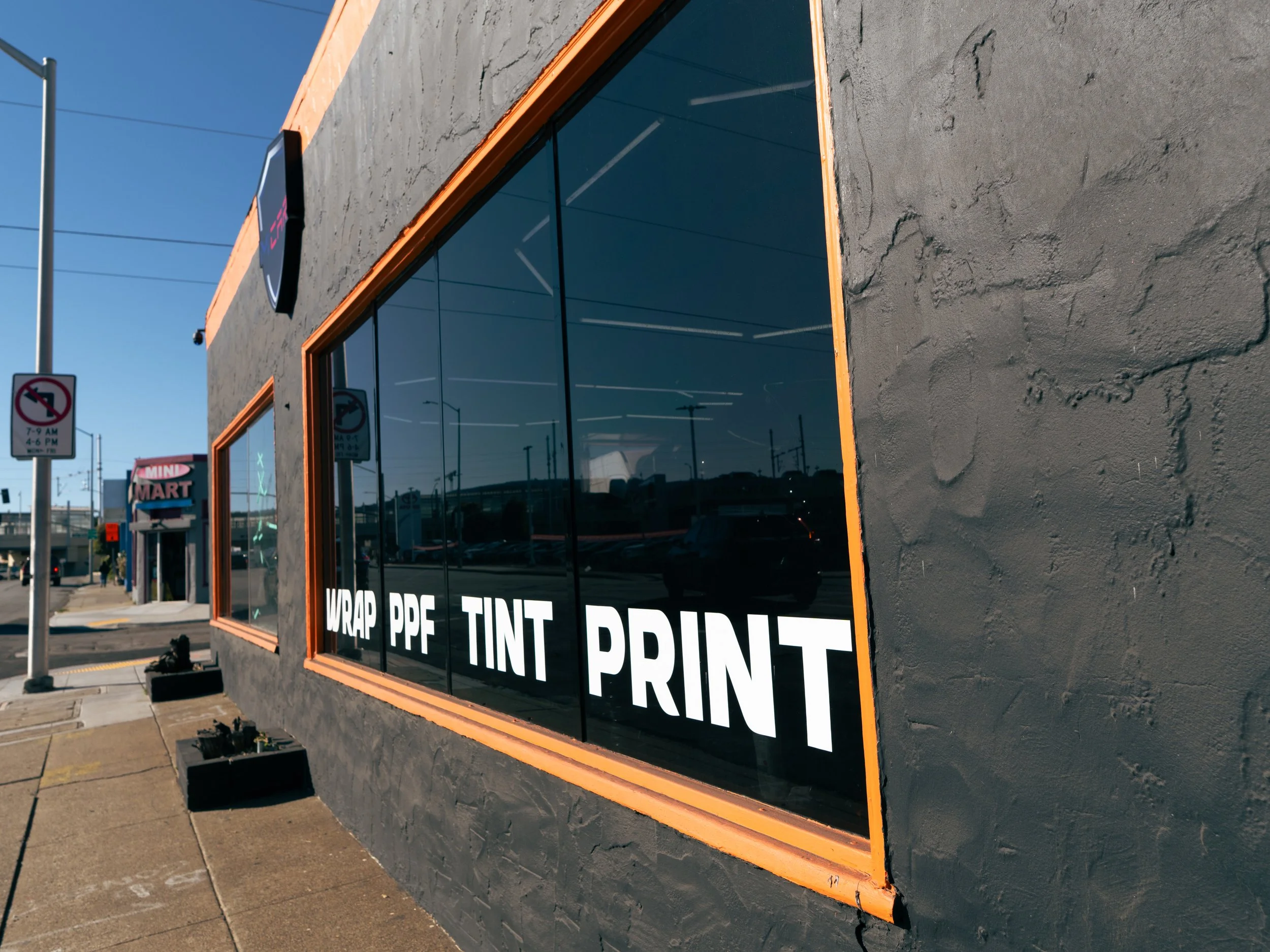 Retail storefront with large tinted windows displaying words: 'WRAP PPF', 'TINT', and 'PRINT' in white letters, with a textured gray wall and orange window frames, located on a sidewalk with parking lot reflections visible in the glass.