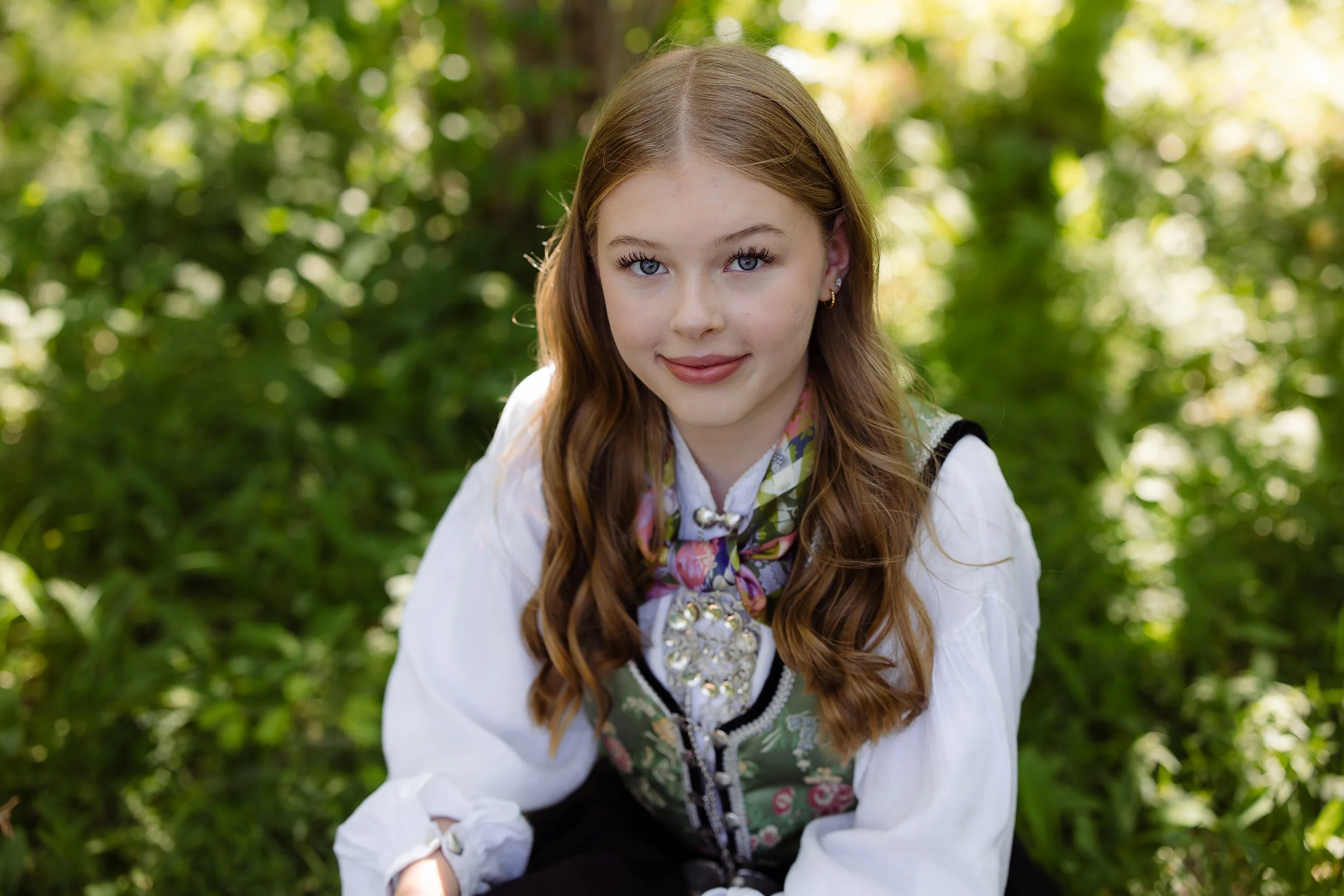 Ung kvinne i tradisjonell norsk bunad, sittende i en grønn skog med sollys. Håret er langt og krøllete, og hun smiler mot kamera.