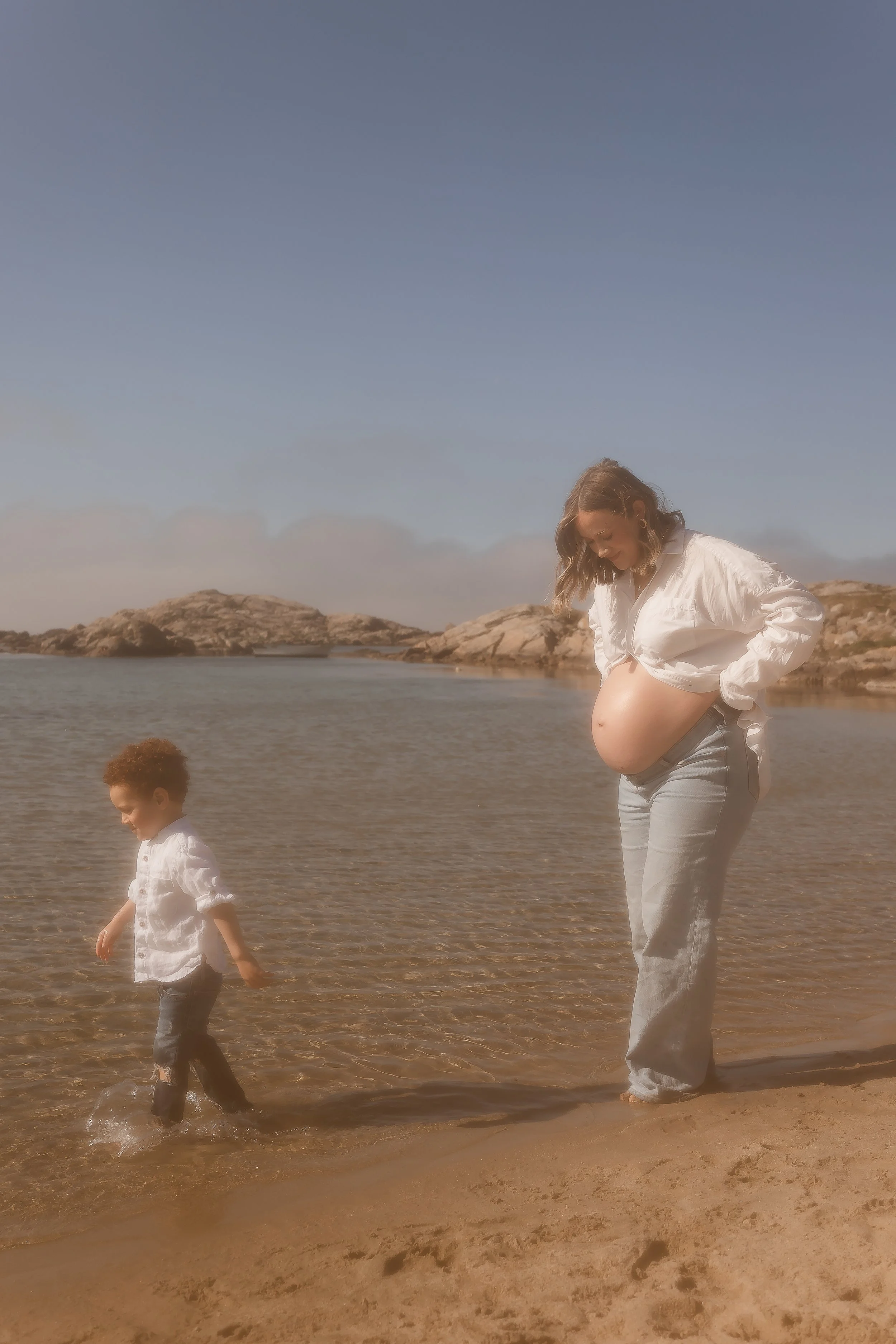 En gravid kvinne og et barn som går langs stranden ved vannkanten, med steinformasjoner i bakgrunnen under en klar himmel.