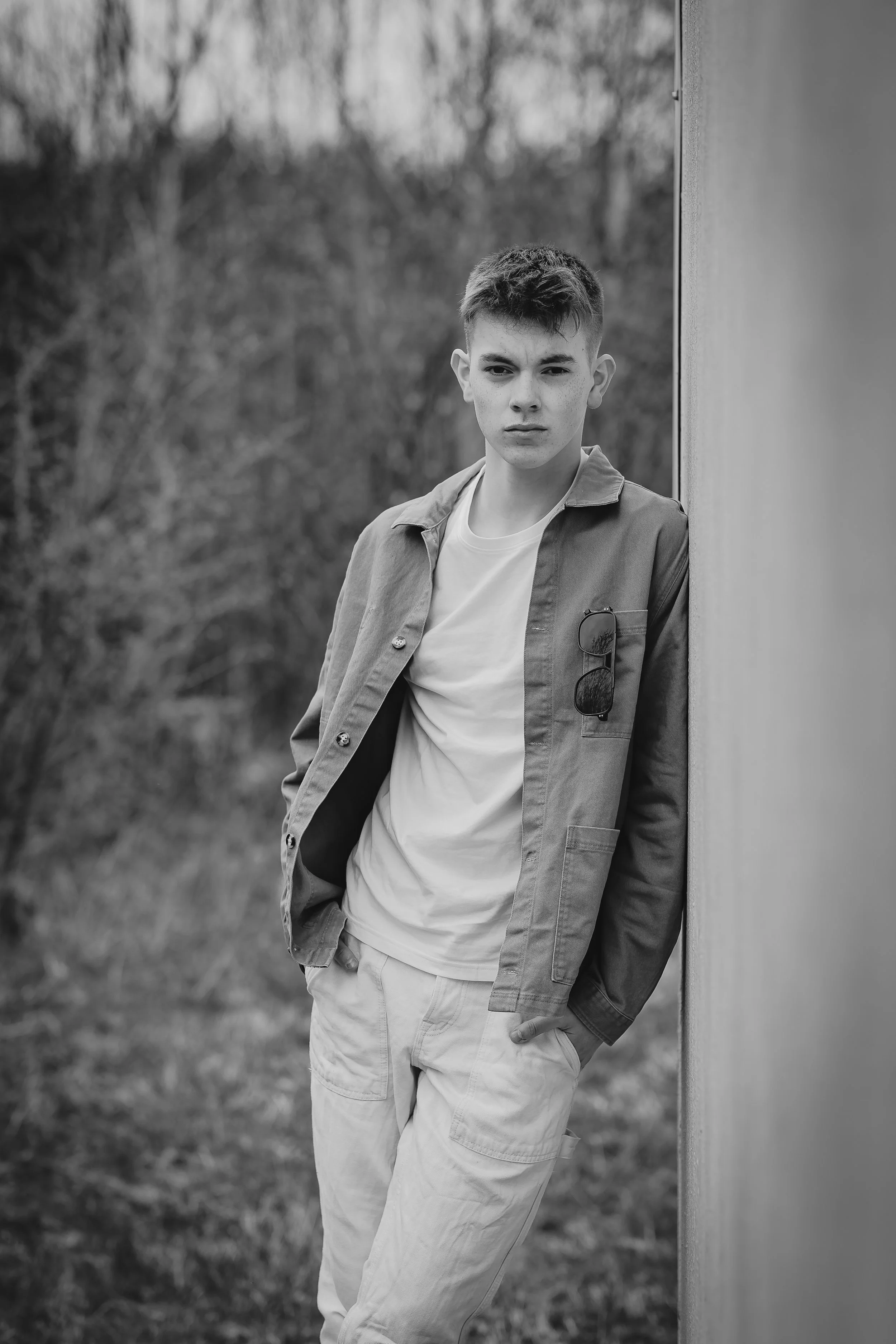 Ung mann står ved en vegg ute i naturen, iført en hvit t-skjorte og en jakke med solbriller hengende på brystet.