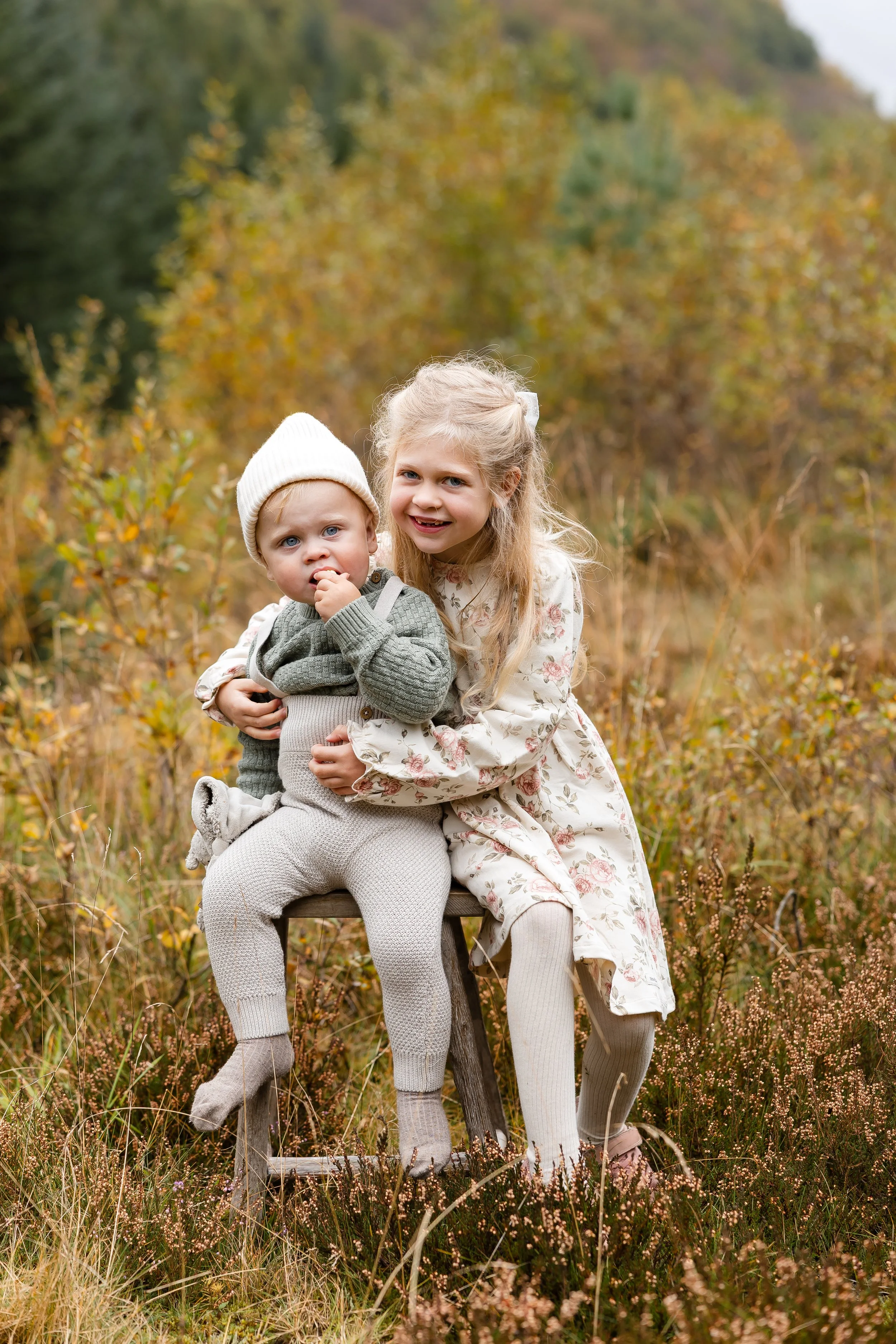To barn, en jente og en yngre gutt, sitter på en trestol i en høstskog, med høstfargede blader i bakgrunnen.