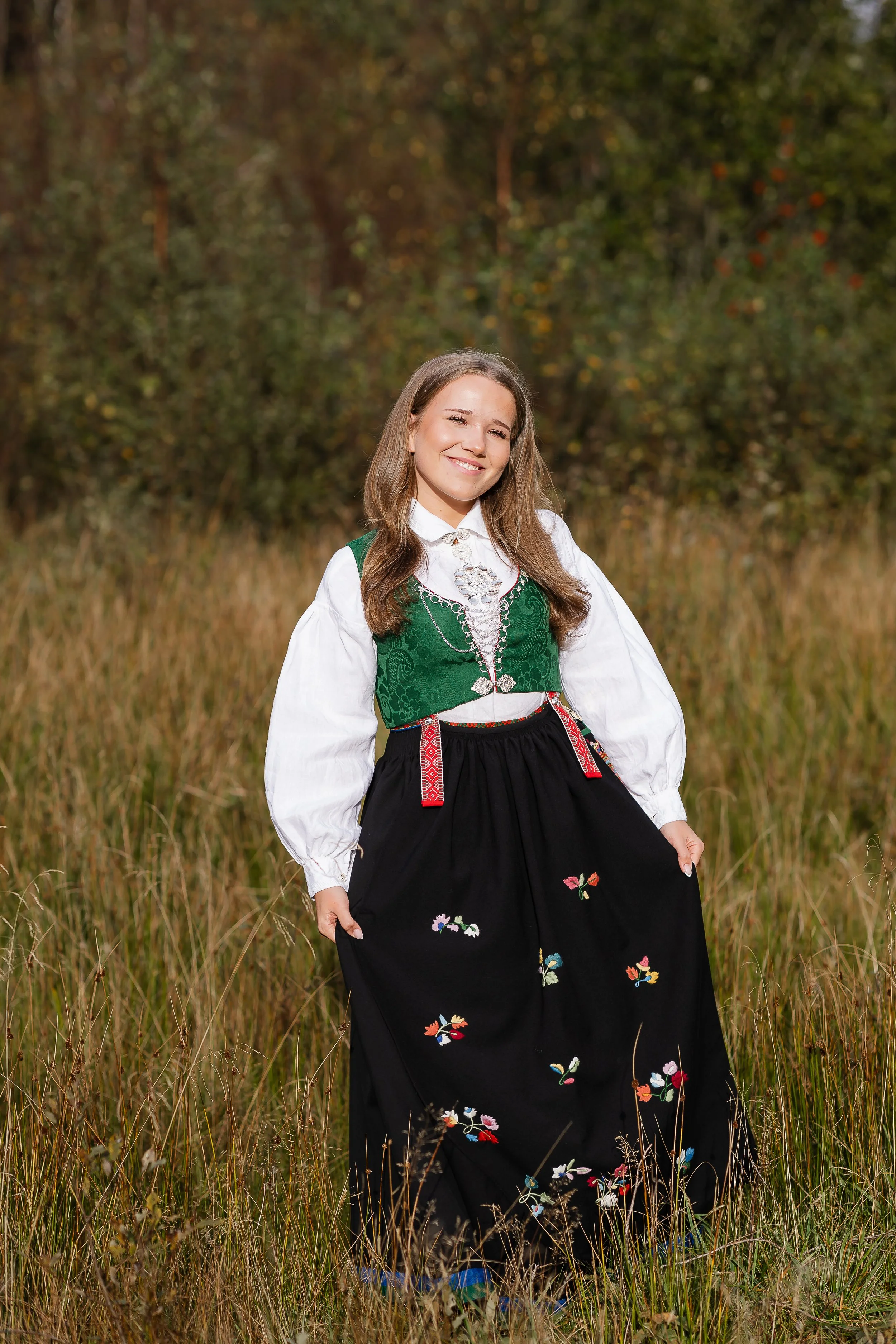 Kvinne i tradisjonell norsk bunad i en åker med høstfarger i bakgrunnen.