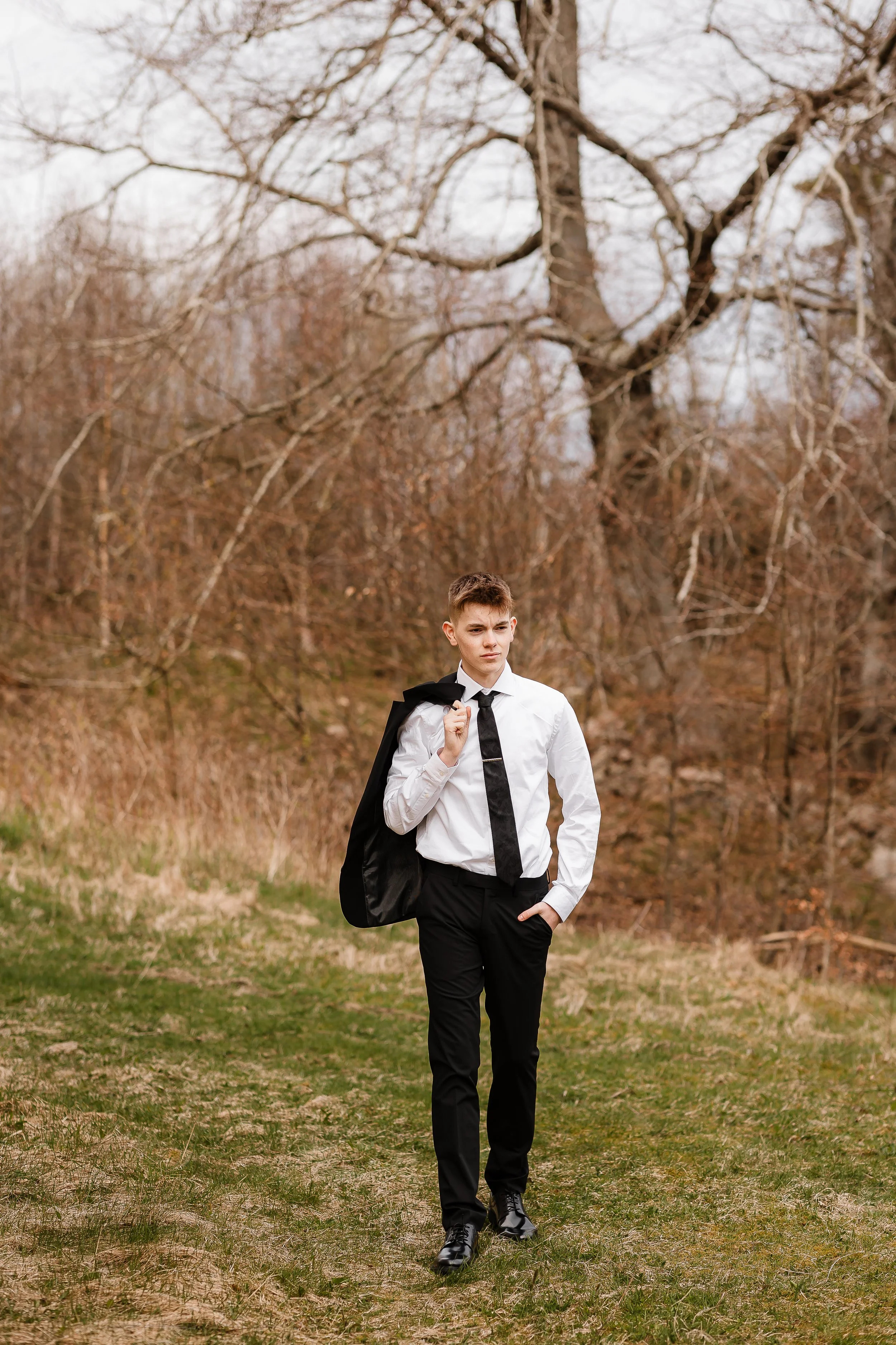 Ung mann i svart dress og hvit skjorte med slips, bærer en svart jakke over skulderen, går på en sti i en skog om høsten.
