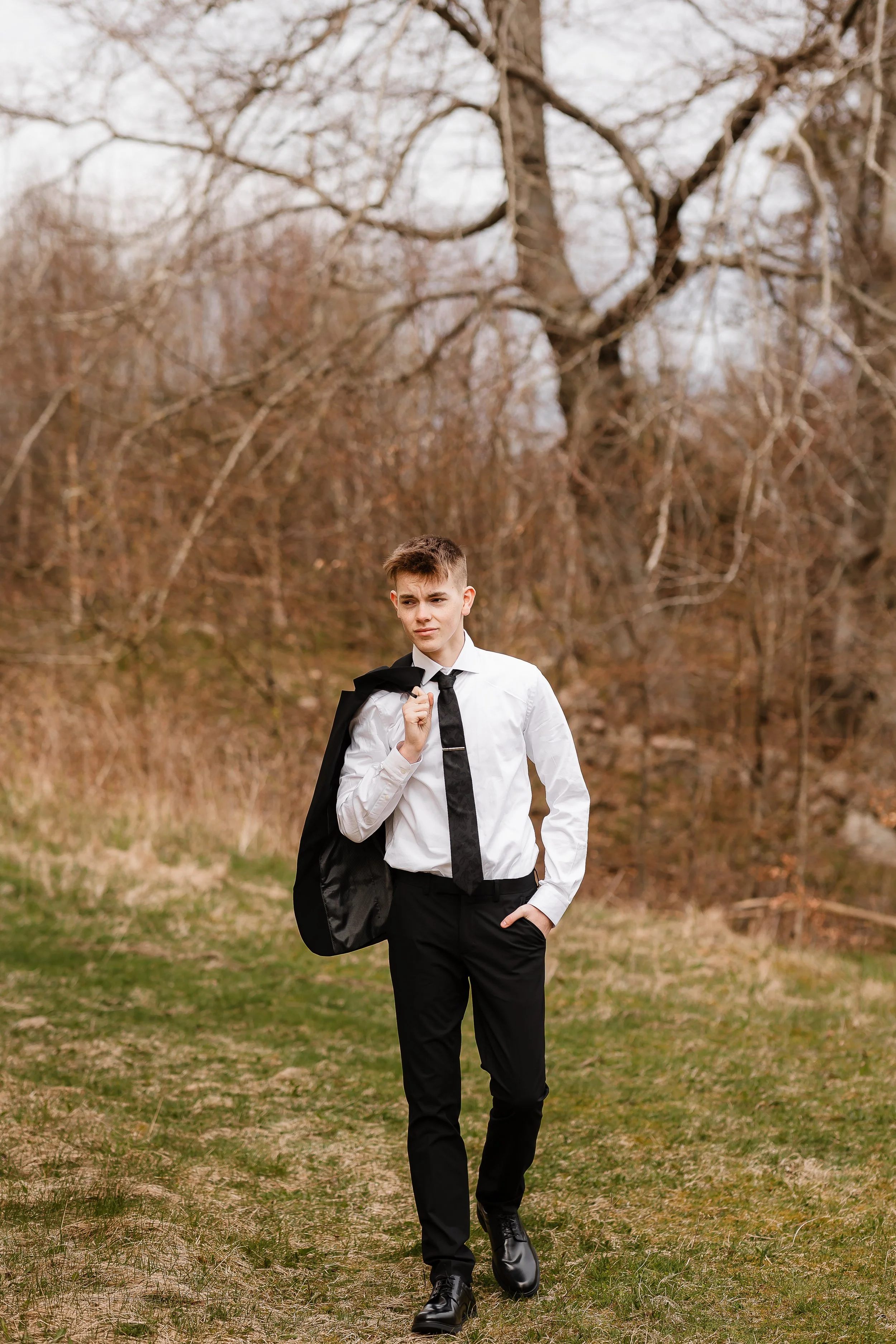 Ung mann i hvit skjorte, svart slips, svarte bukser, og sorte sko går på en sti i en skog. Han holder en svart jakke over skulderen med en hånd og har den andre i lommen. Bakgrunnen består av løvtrær uten blader.