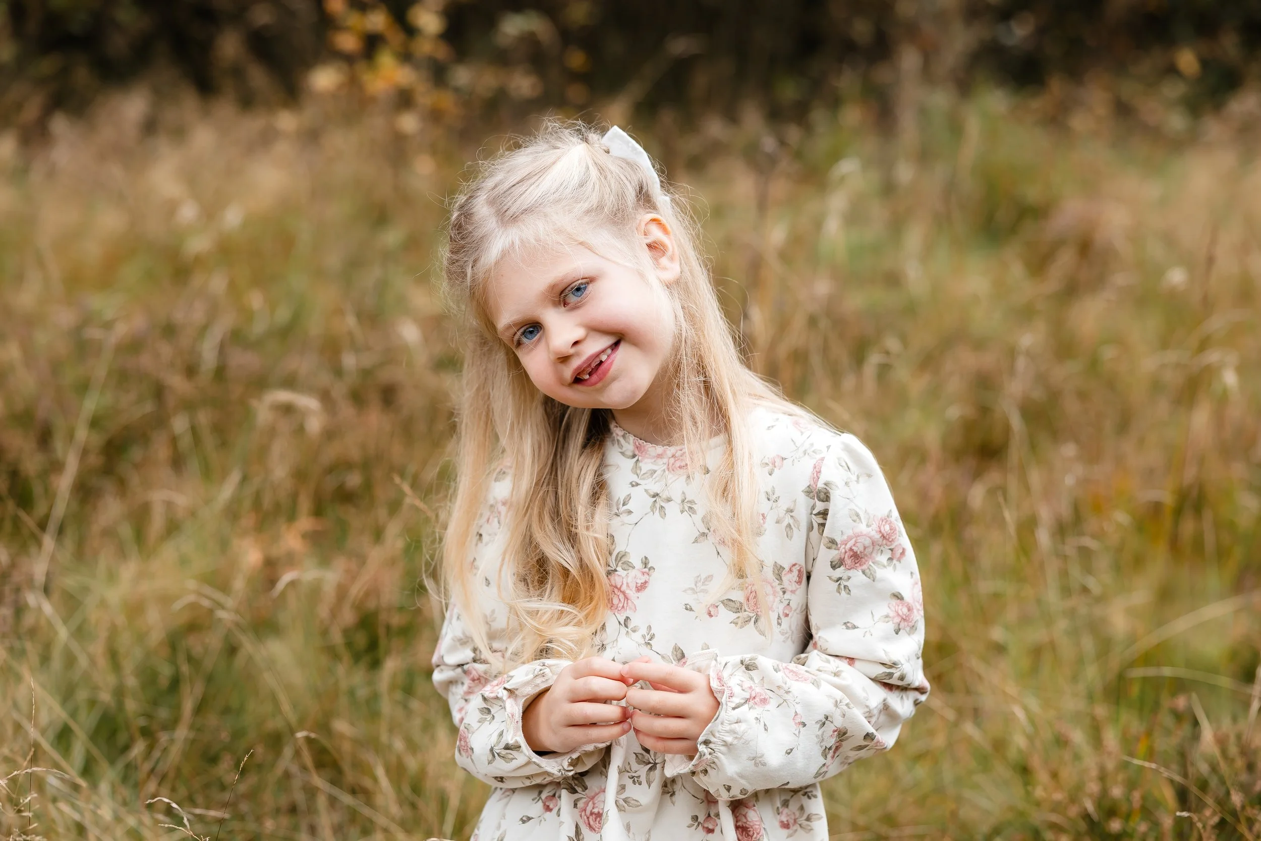 En smilende ung jente med langt blondt hår og blå øyne, kledd i en hvit kjole med blomster, står i et gresslette med brunt treverk i bakgrunnen.