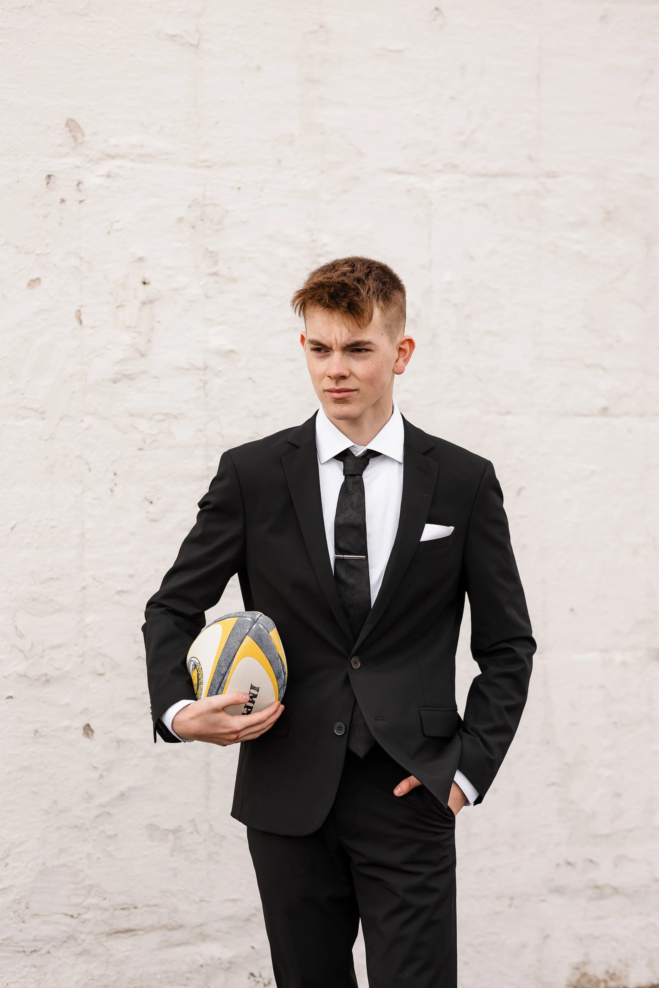 Ung mann i svart dress og slips holder en rugbyball mot en hvit murvegg.