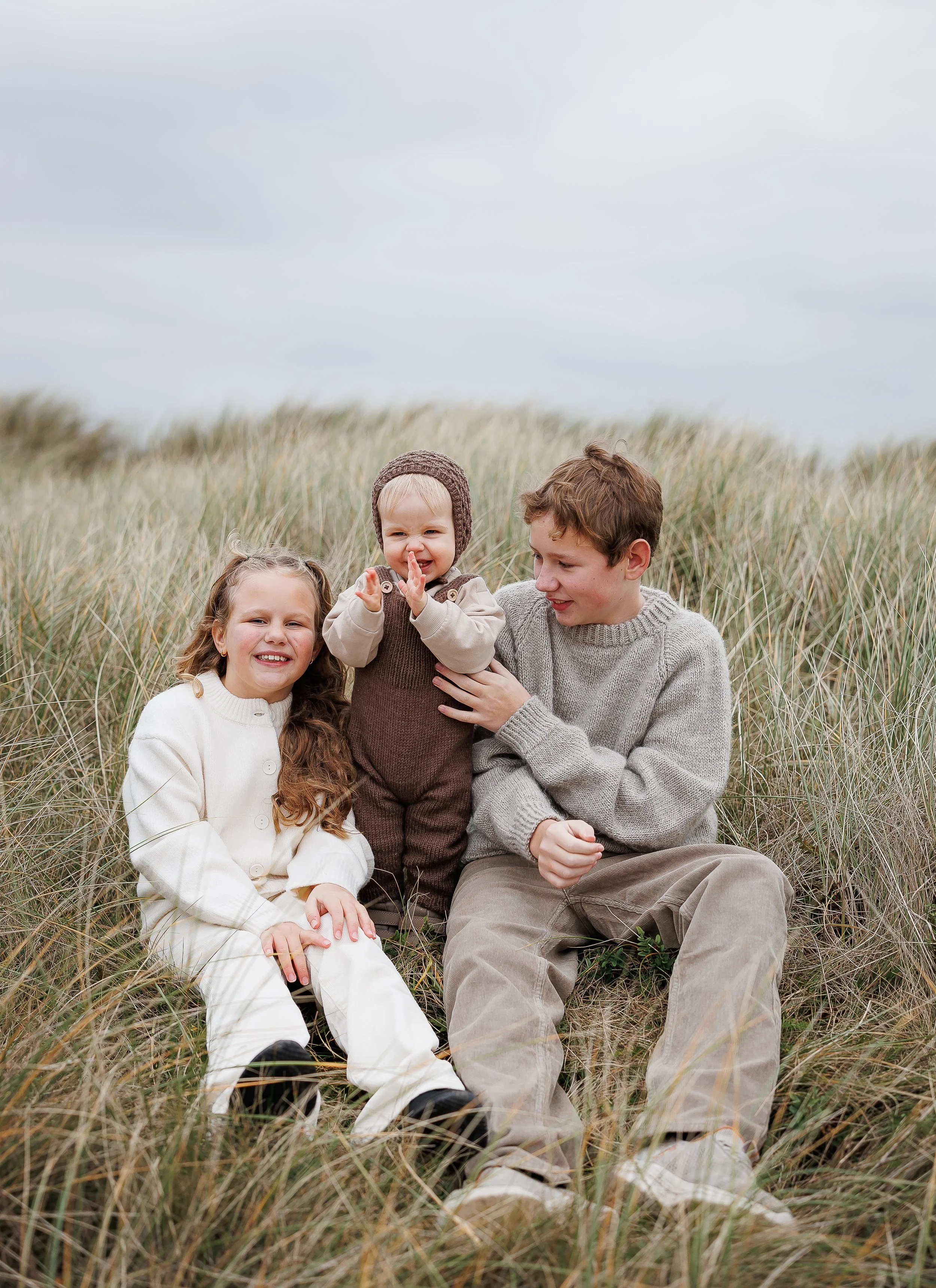 Tre barn som sitter i høyt gress på en ås, smiler og leker sammen, med en overskyet himmel i bakgrunnen.