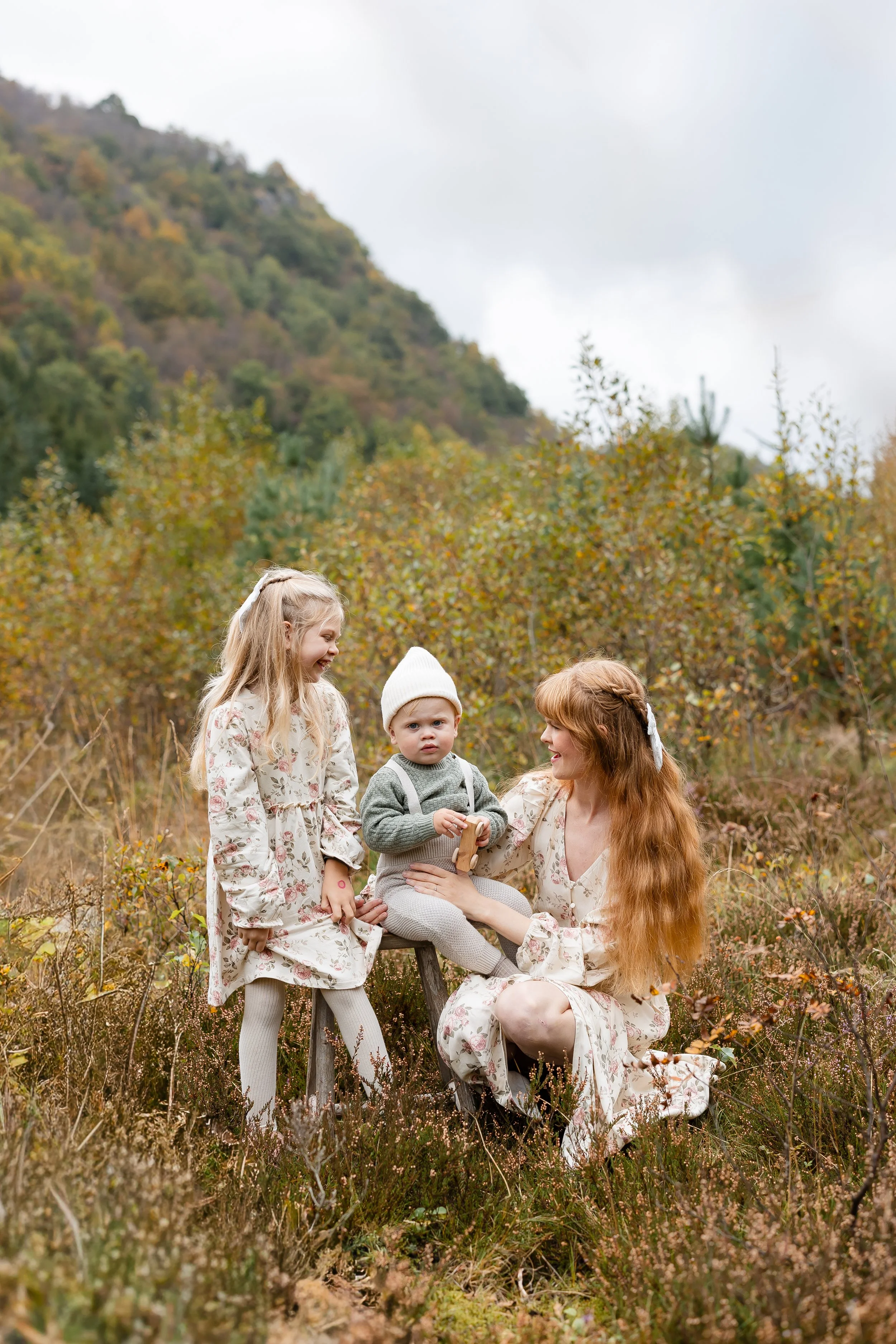En kvinne med lang rød hår og to unge jenter i en høstskog, alle iført blomsterkjoler, med ett barn sittende på en liten stol og det andre ved siden av.