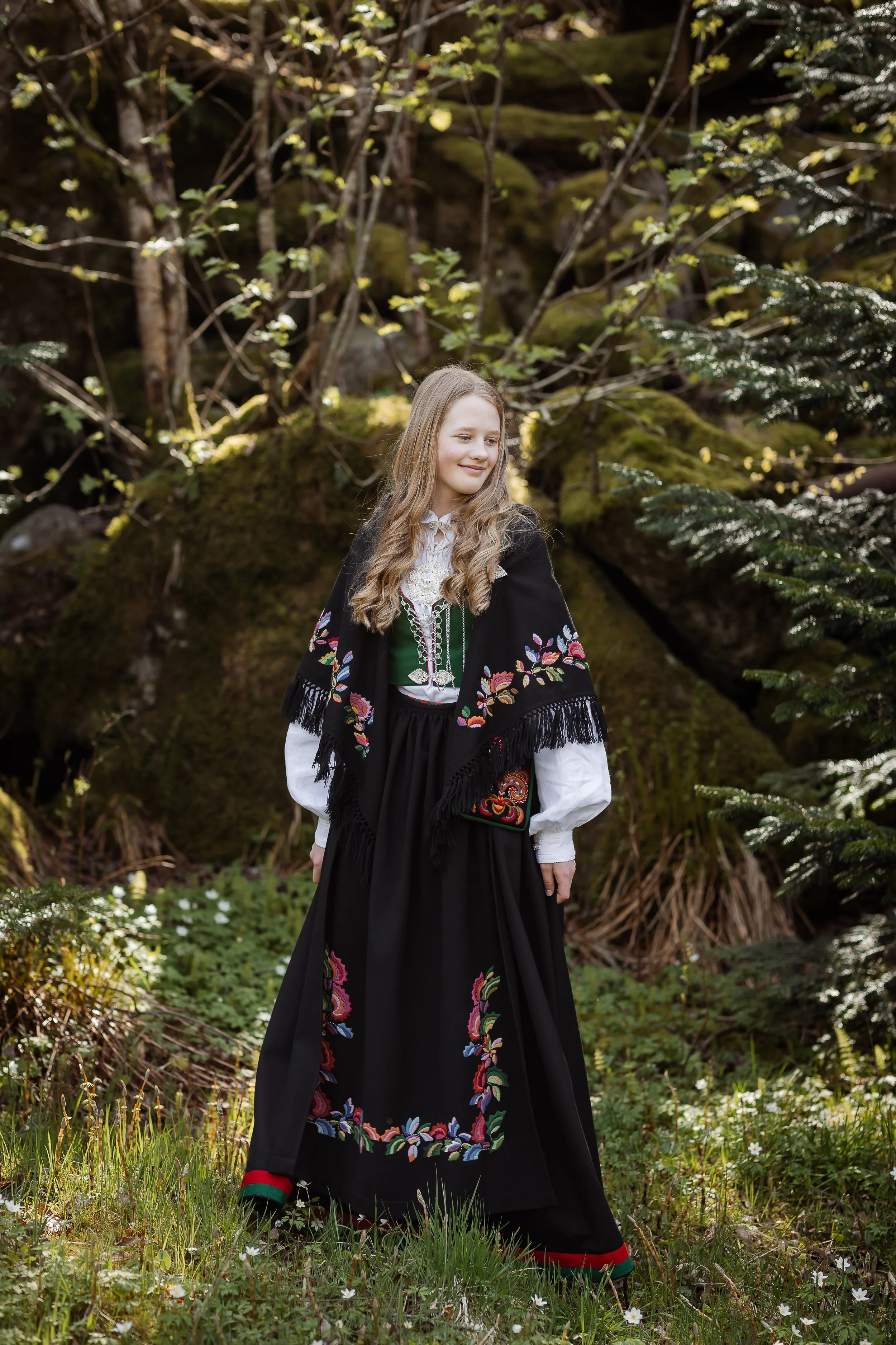 En ung jente som står i skogen iført tradisjonell norsk folkdrakt med blomsterbroderier. Hun har langt, lyst hår og smiler svakt.