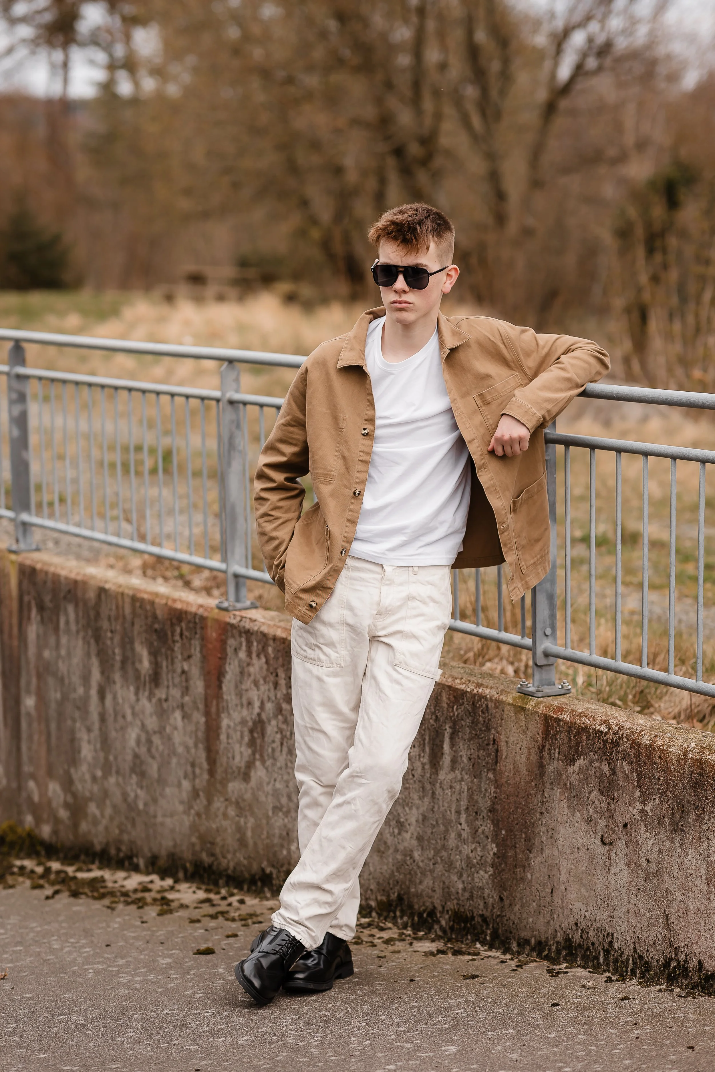 Ung mann med solbriller, brun jakke, hvit t-skjorte, lyse bukser og svarte støvler står ved en metallbarriere på en grusvei, omgitt av høstfarger trær.
