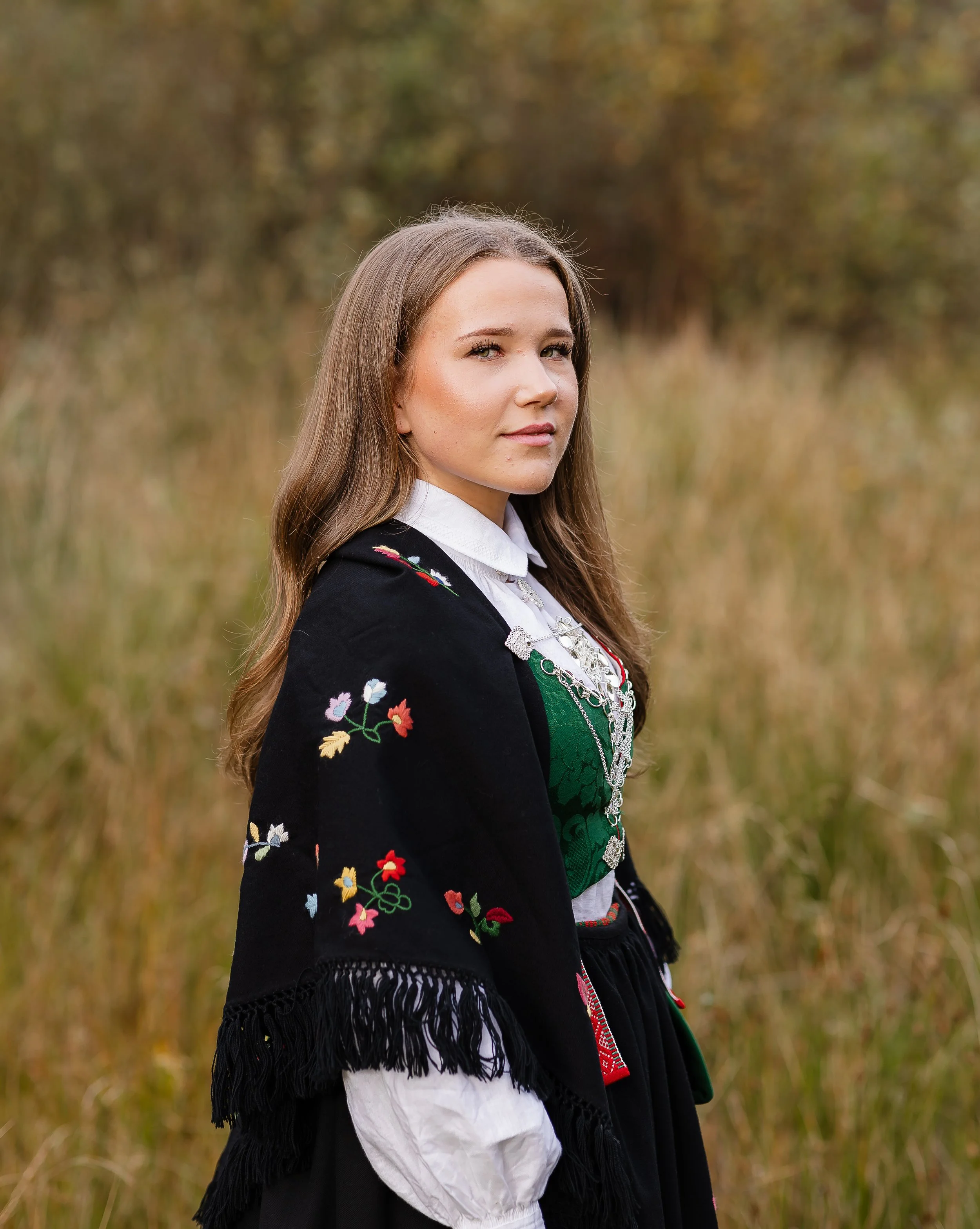 En ung kvinne i tradisjonell norsk folkedrakt står i et åpent høstlandskap.