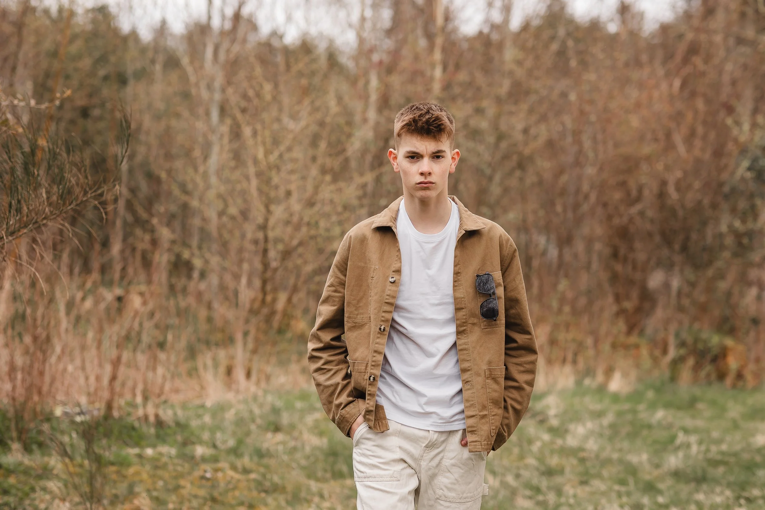 En ung mann med kort, brunt hår står i en veikant med bartrær og busker i bakgrunnen, iført en brunslitt jakke, hvit t-skjorte og beige bukser.