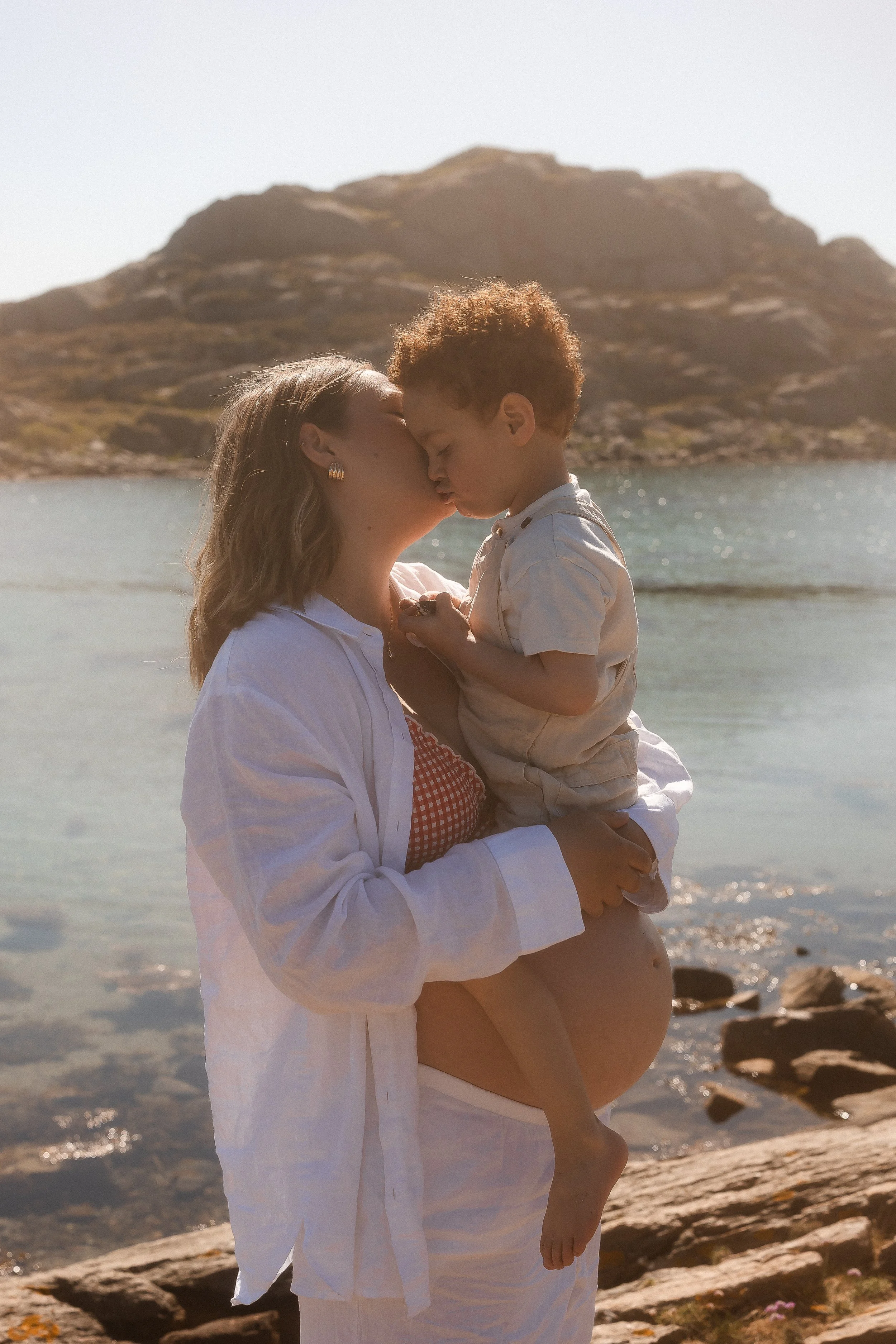 En kvinne holder en liten gutt, de kysser hverandre foran havet og en fjellkjede i bakgrunnen på en solrik dag.