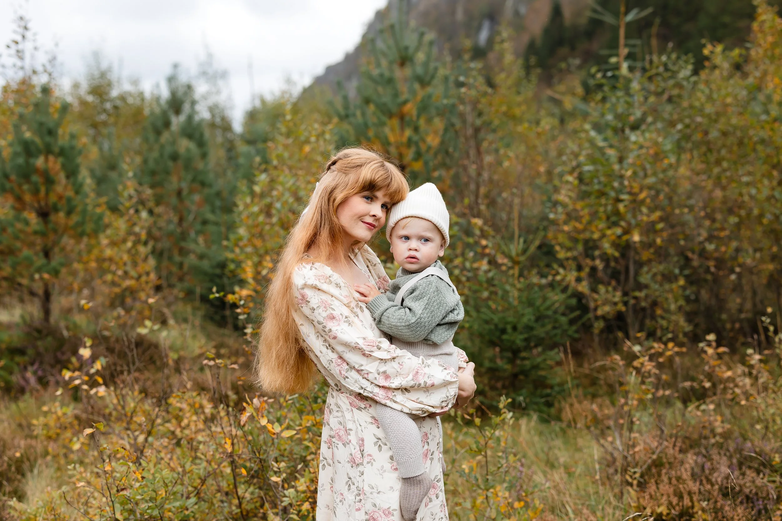 En kvinne med langt, lyst hår holder en liten gutt i en skog med høstfargede trær.