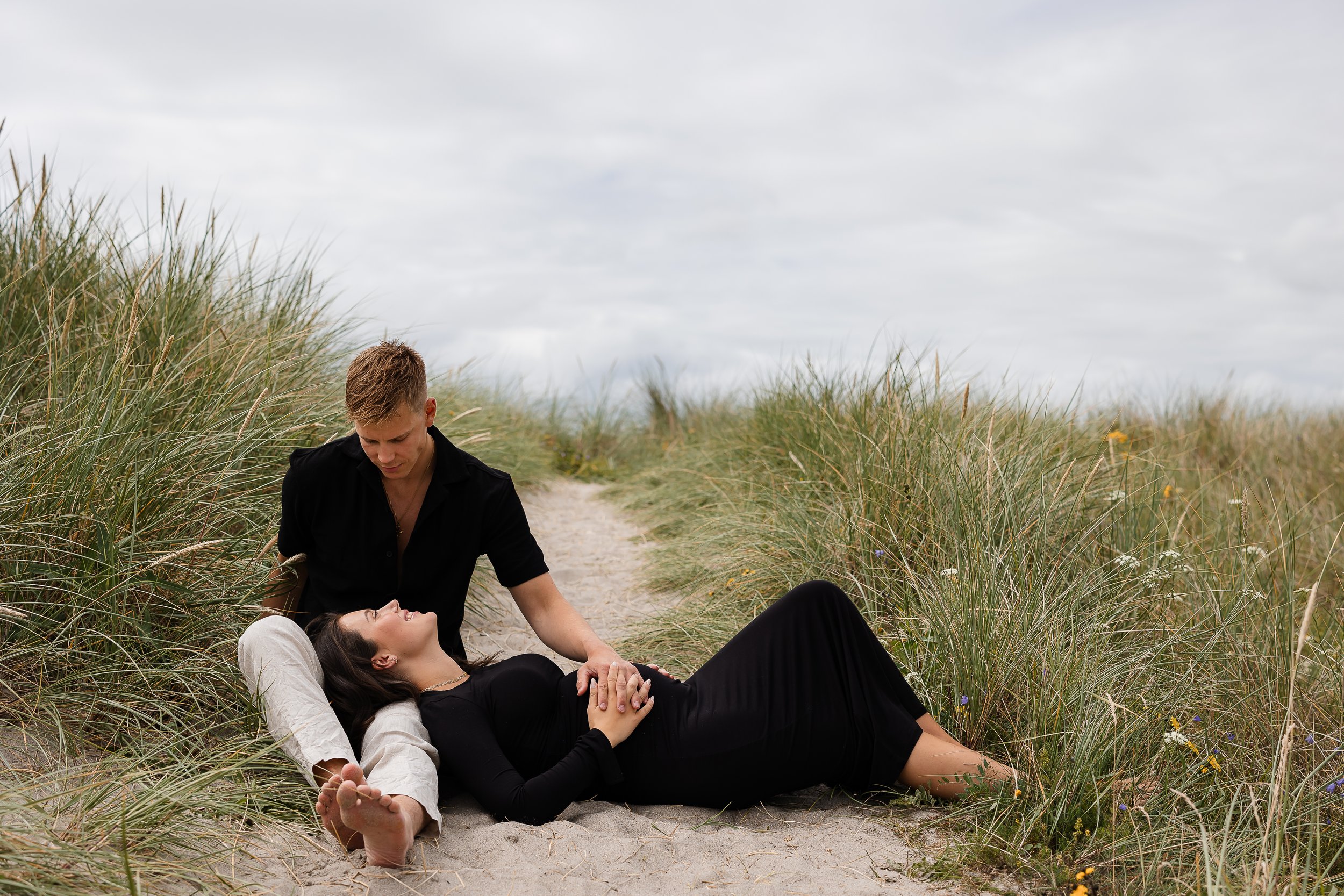 En mann og kvinne ligger på sand mellom gresskledde sanddyner på en overskyet dag, der kvinnen ligger ned og mannen sitter ved siden av henne, holder hender og ser ned mot henne.