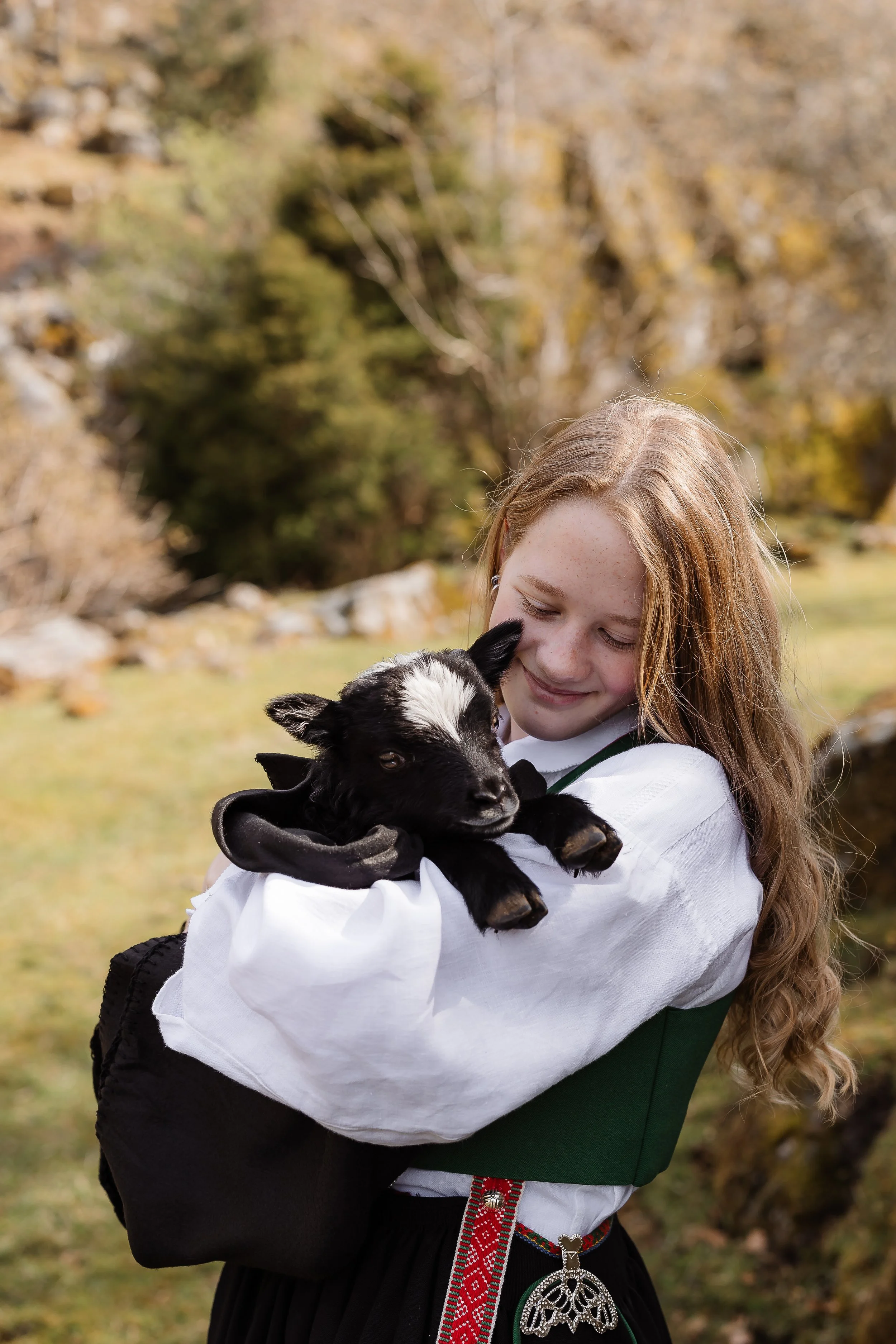 Jente i tradisjonell norsk bunad holder en liten svart og hvit geitebukk i fanget.