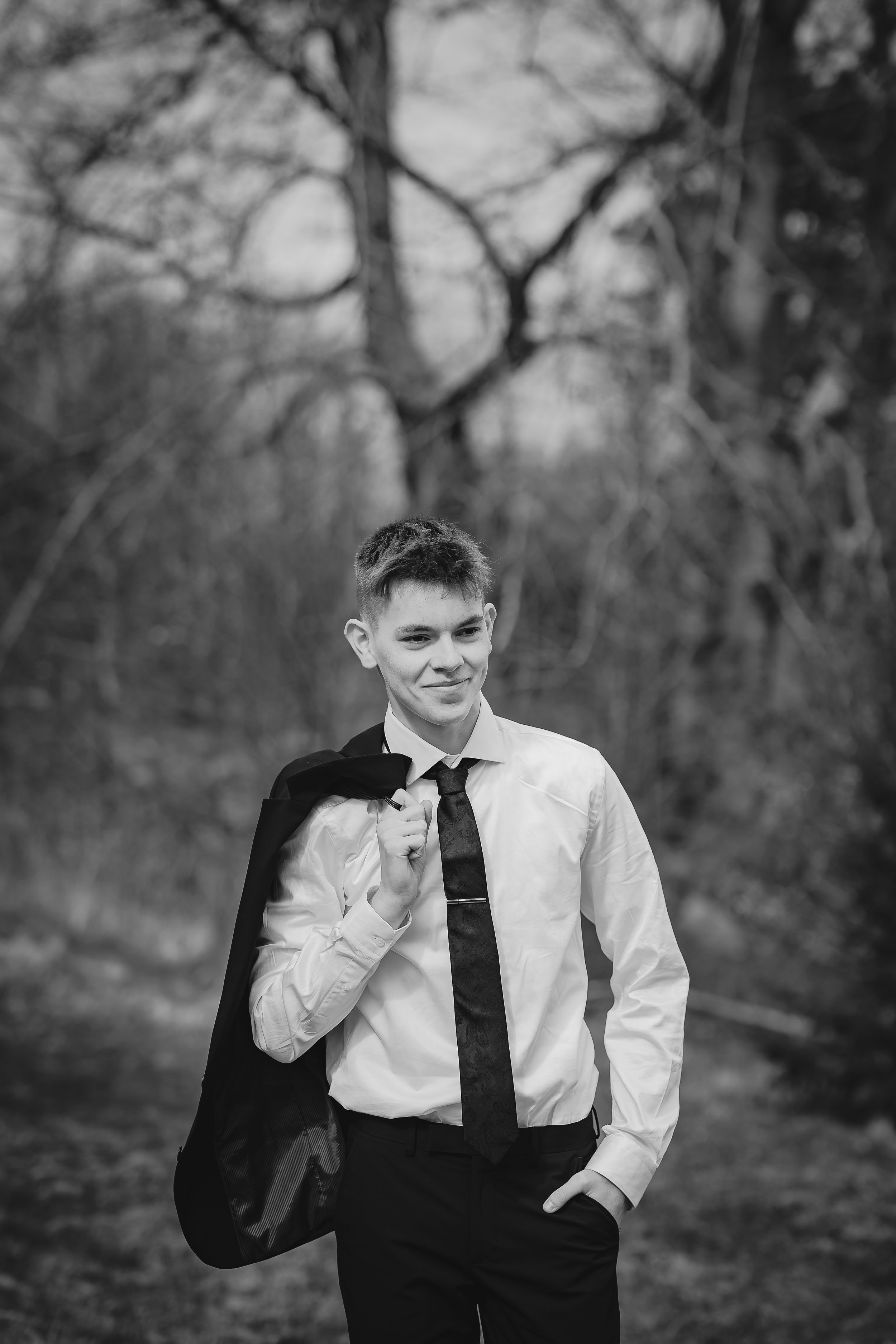 En ung mann i hvit skjorte og slips går utendørs med en svart jakke over skuldrene og en svart veske på armen, omgitt av trær uten løv. Bildet er i svart-hvitt.