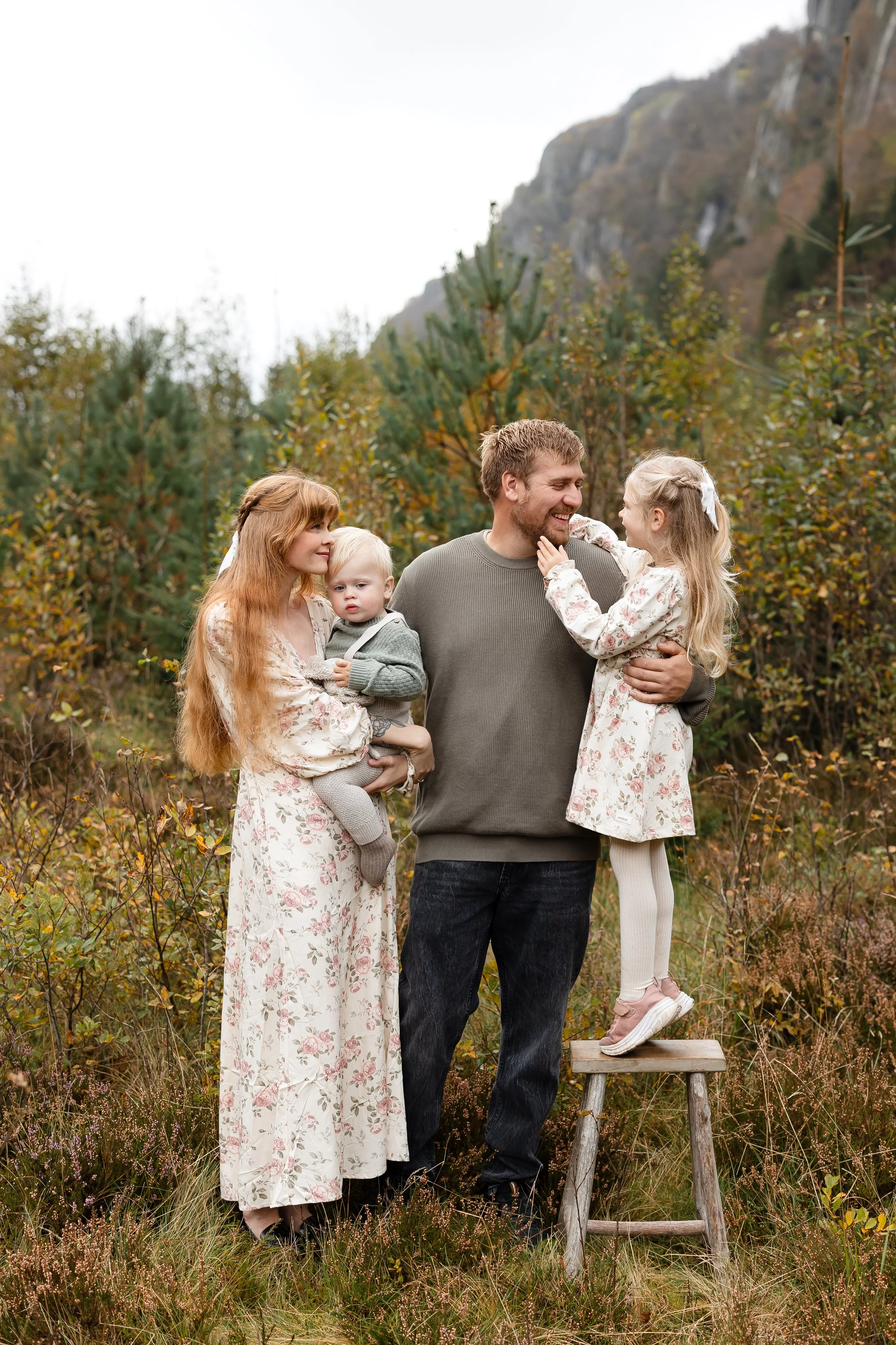 Familie på fire i en skog med høstfarger, man holder en datter på armen, kvinnen holder et barn, og mannen står i midten med datteren som står på en stol.