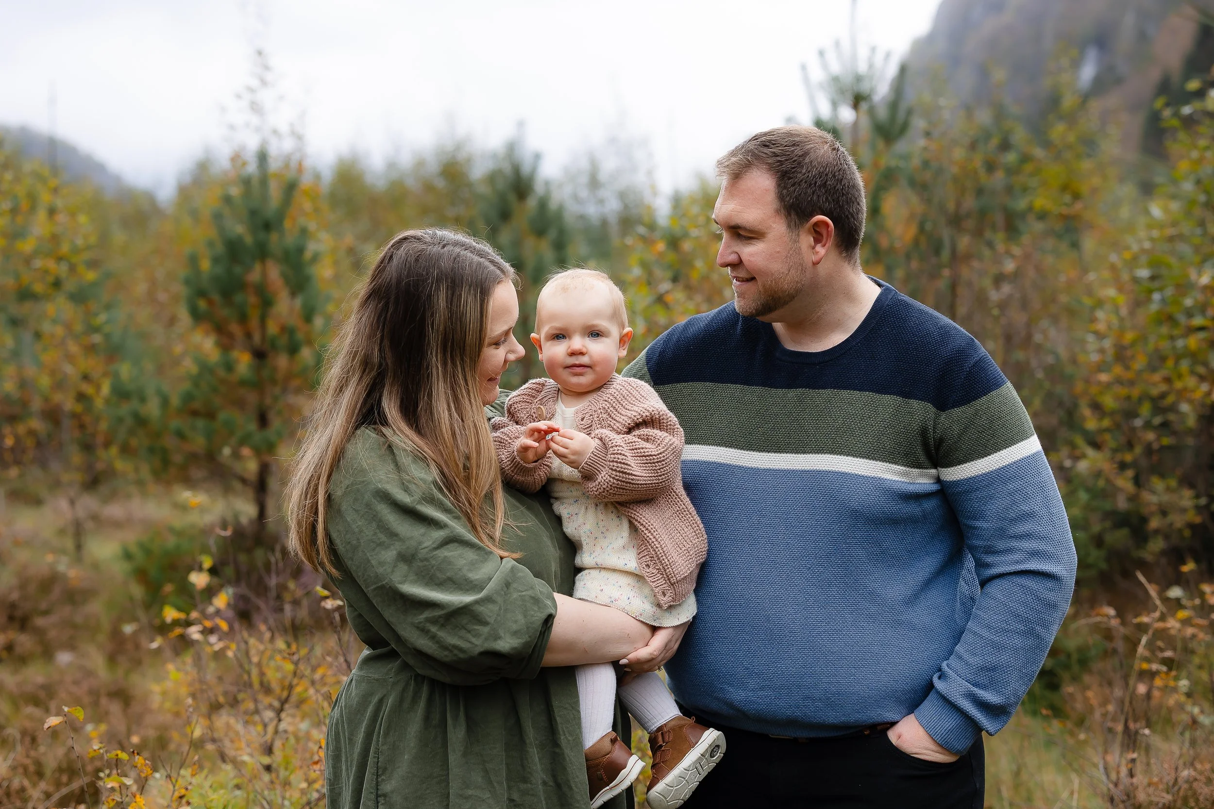 En familie på tre står ute i naturen, med høstfarger på trærne. En kvinne holder en liten jente, mens en mann står ved siden av, alle smiler.