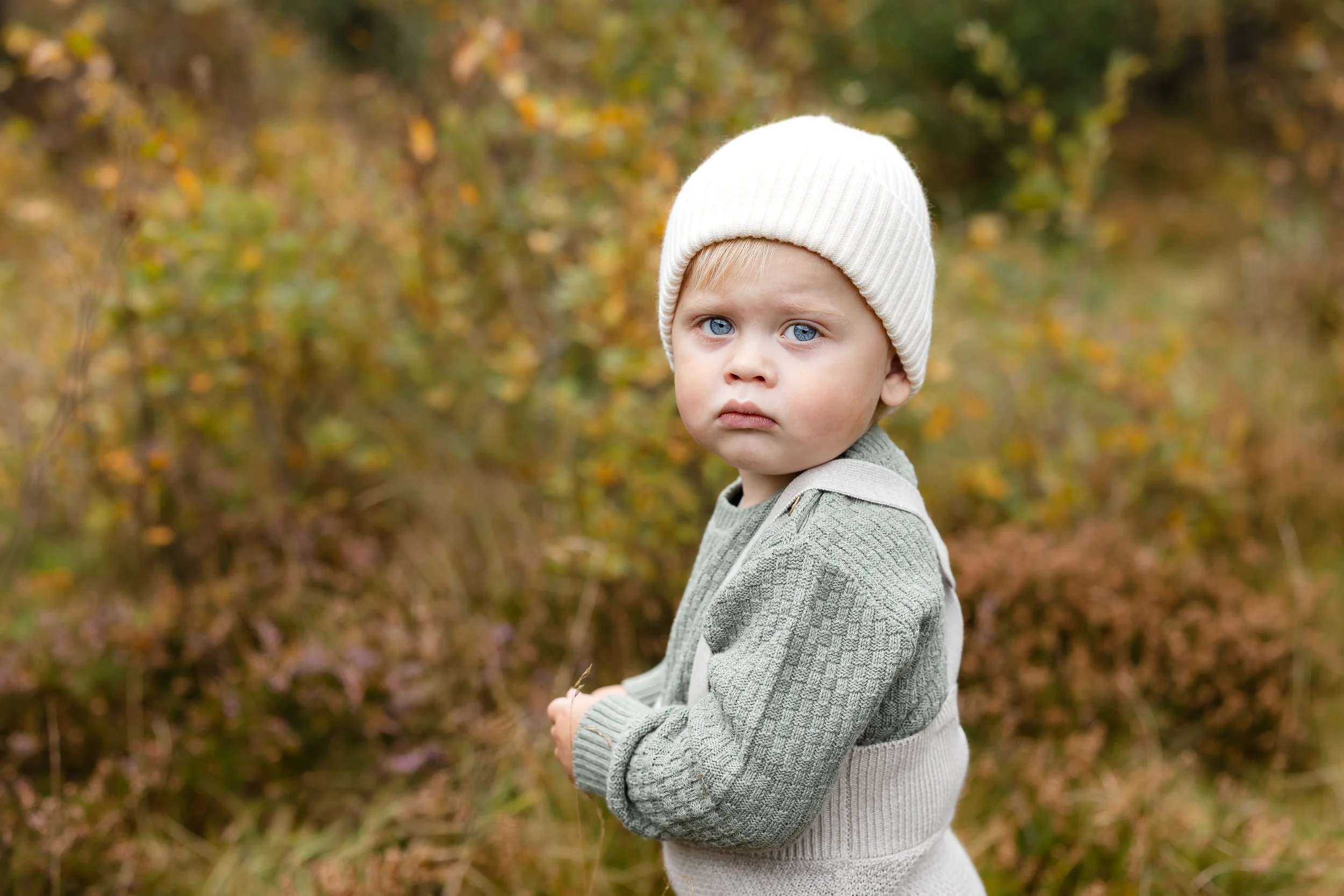 En liten gutt med blå øyne, grå genser og hvit lue står i en skog med høstfarger.