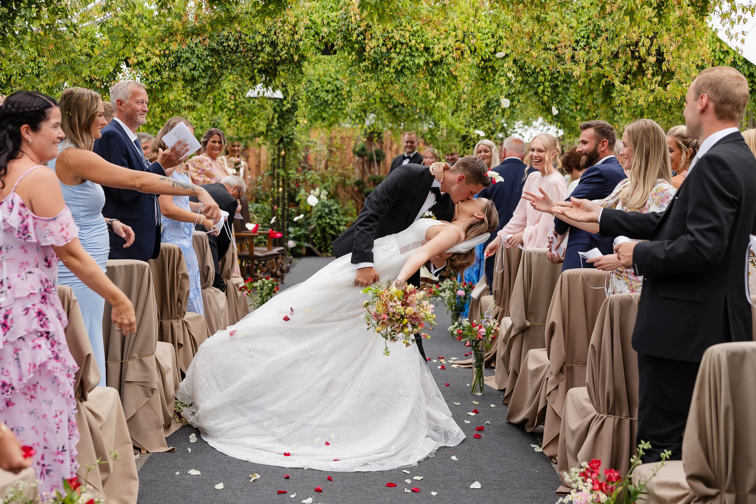 Brudepar som kysser under vielsen, omgitt av venner og familie, i en utendørs hage med grønne trær og blomster.