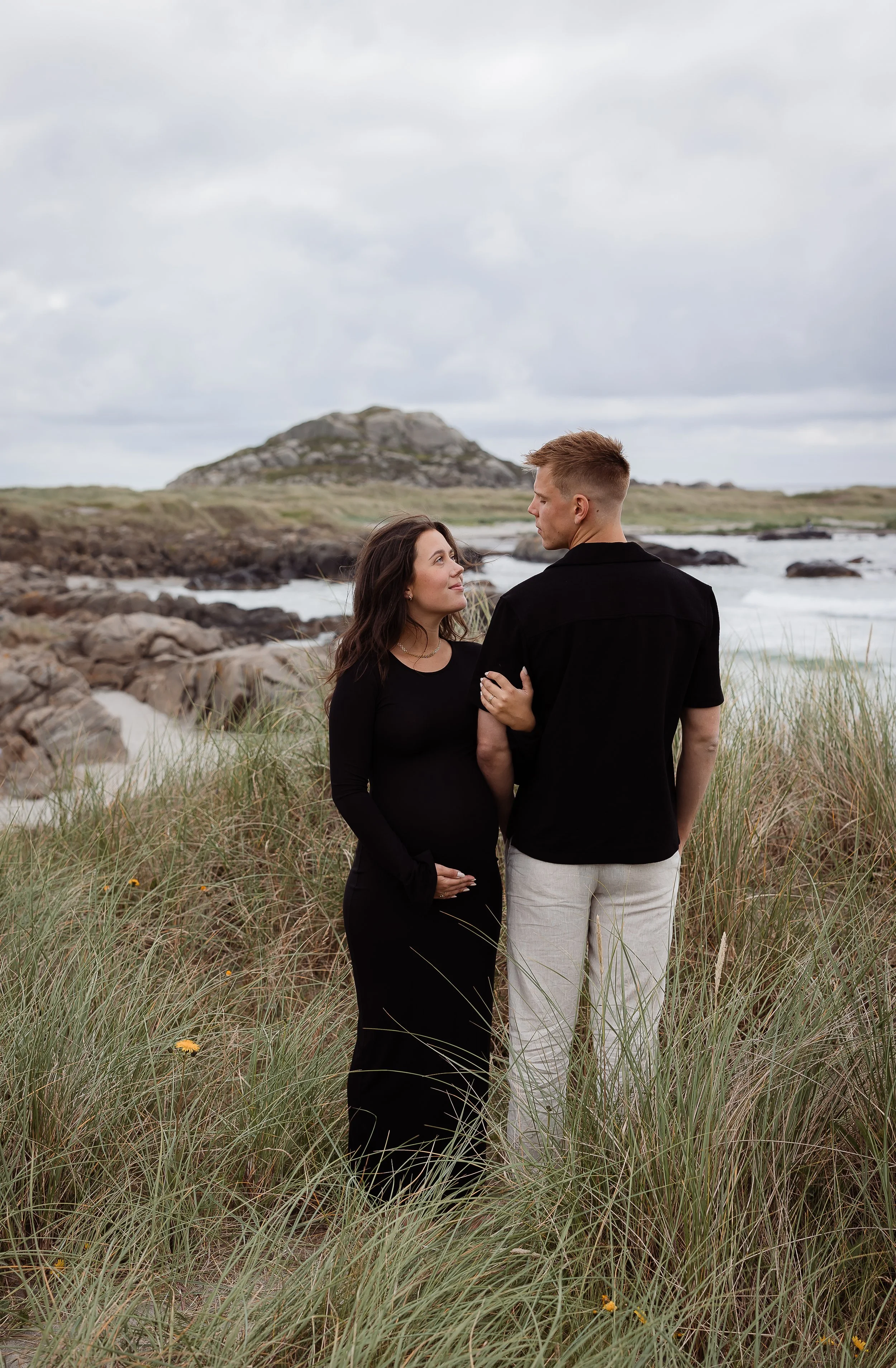 Et par står i en grønn, urteaktig kystlandskap omringet av høye gress, med hav og en øy i bakgrunnen, under overskyet himmel.