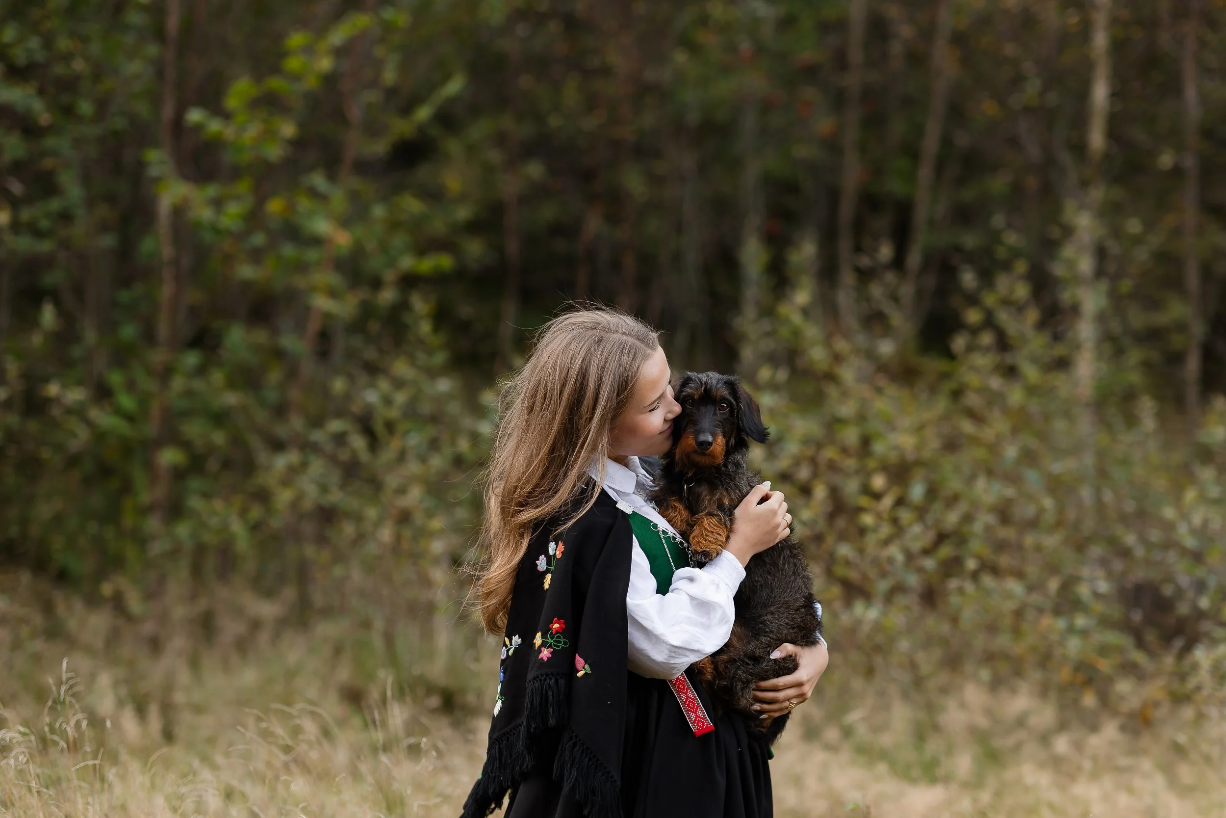 En jente i tradisjonell norsk bunad holder en schäferhund i skogen om høsten.