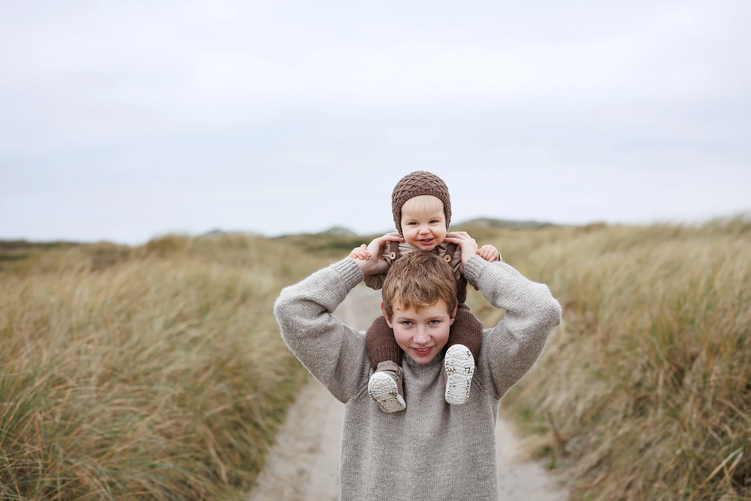 En eldre gutt bærer en liten baby på skuldrene på en sti omgitt av gress. Begge smiler.