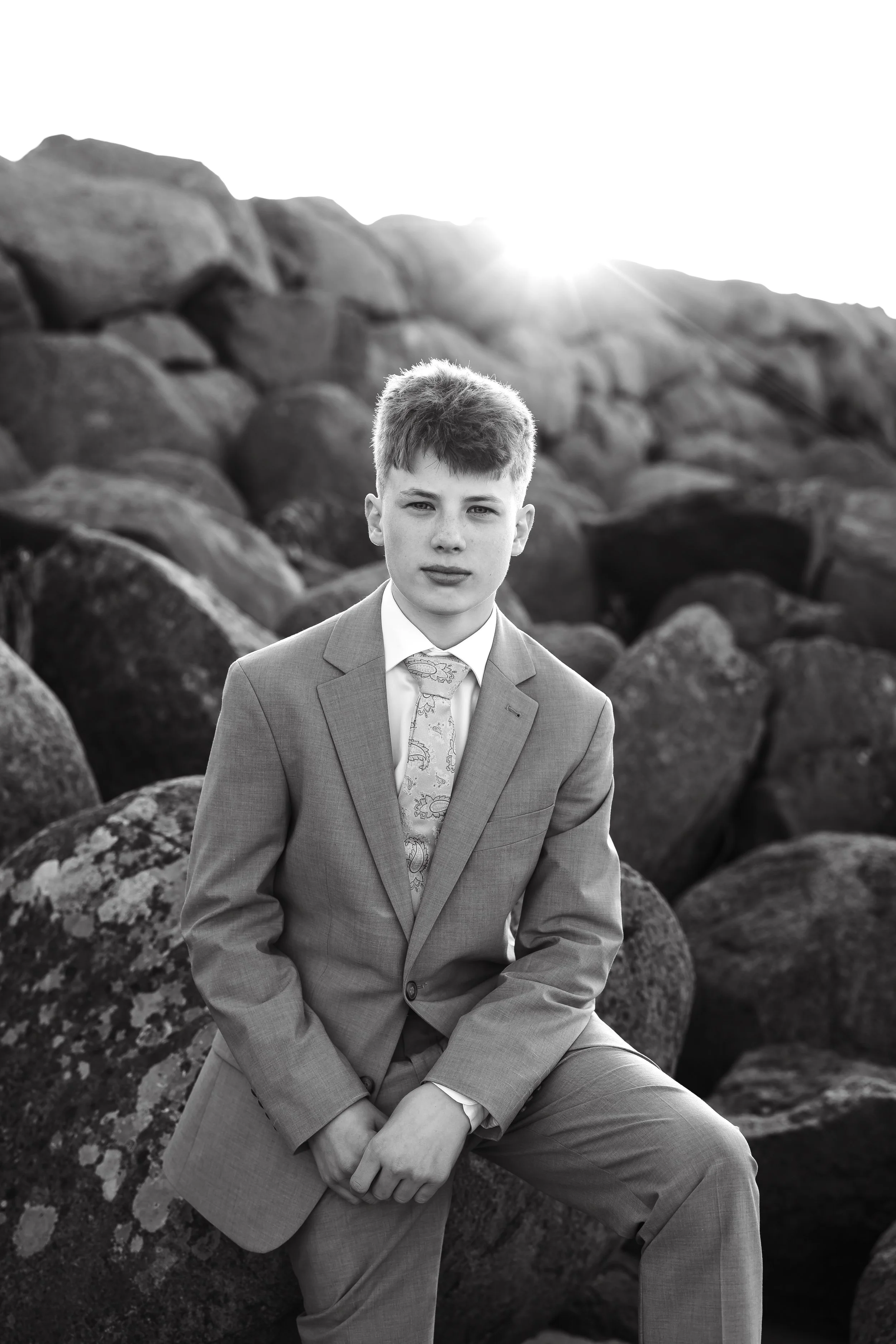 En ung mann i dress sitter på store steiner med fjell i bakgrunnen, fotografert i sort-hvitt.