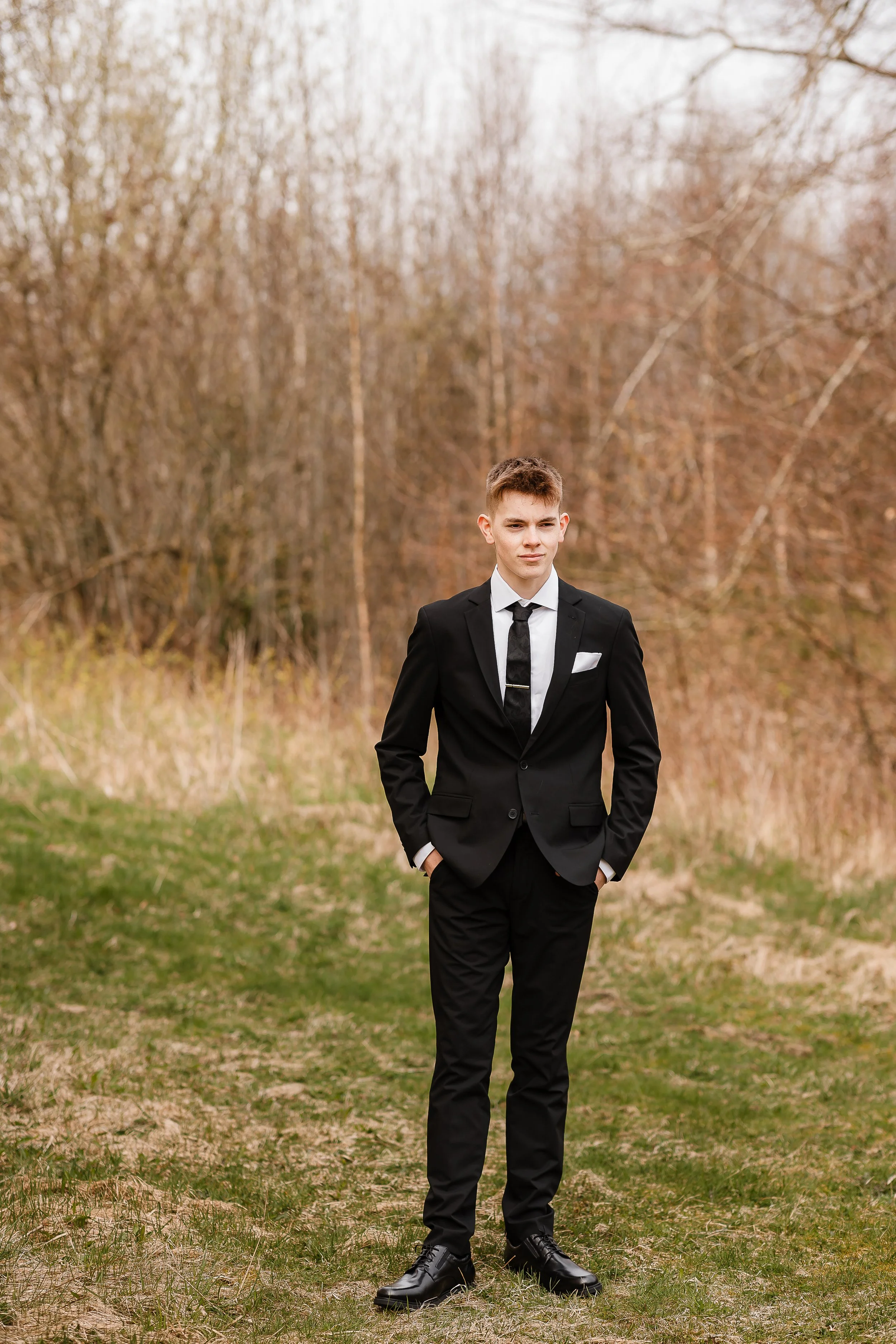 En ung mann i svart dress, hvit skjorte og svart slips står ute i en natur med trær uten blader, sannsynligvis høst eller tidlig vår.