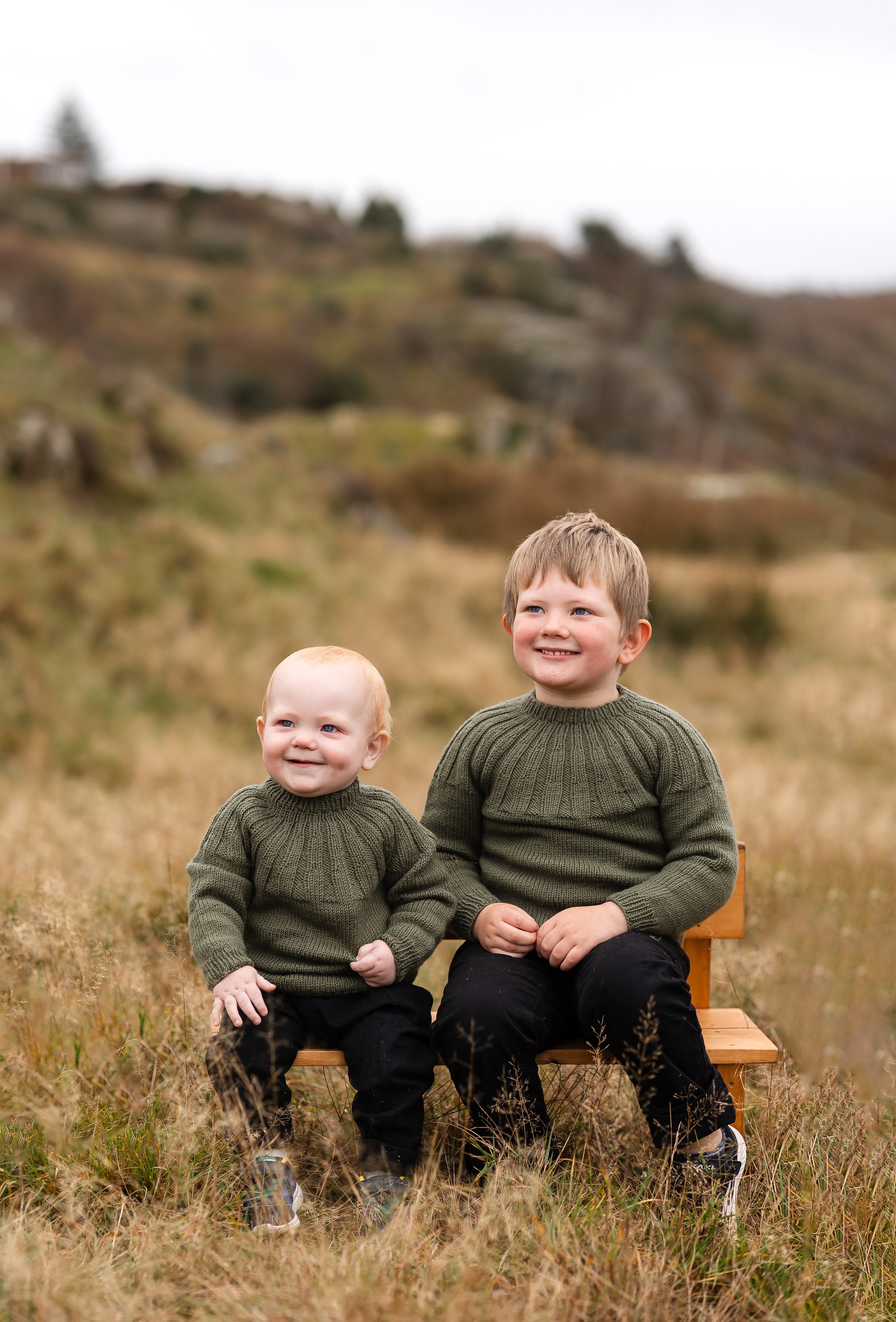 To barn sitter på en trebenk i et åkerlandskap med høstfarger, iført matchende grønne gensere, smilende og ser opp mot høyre.